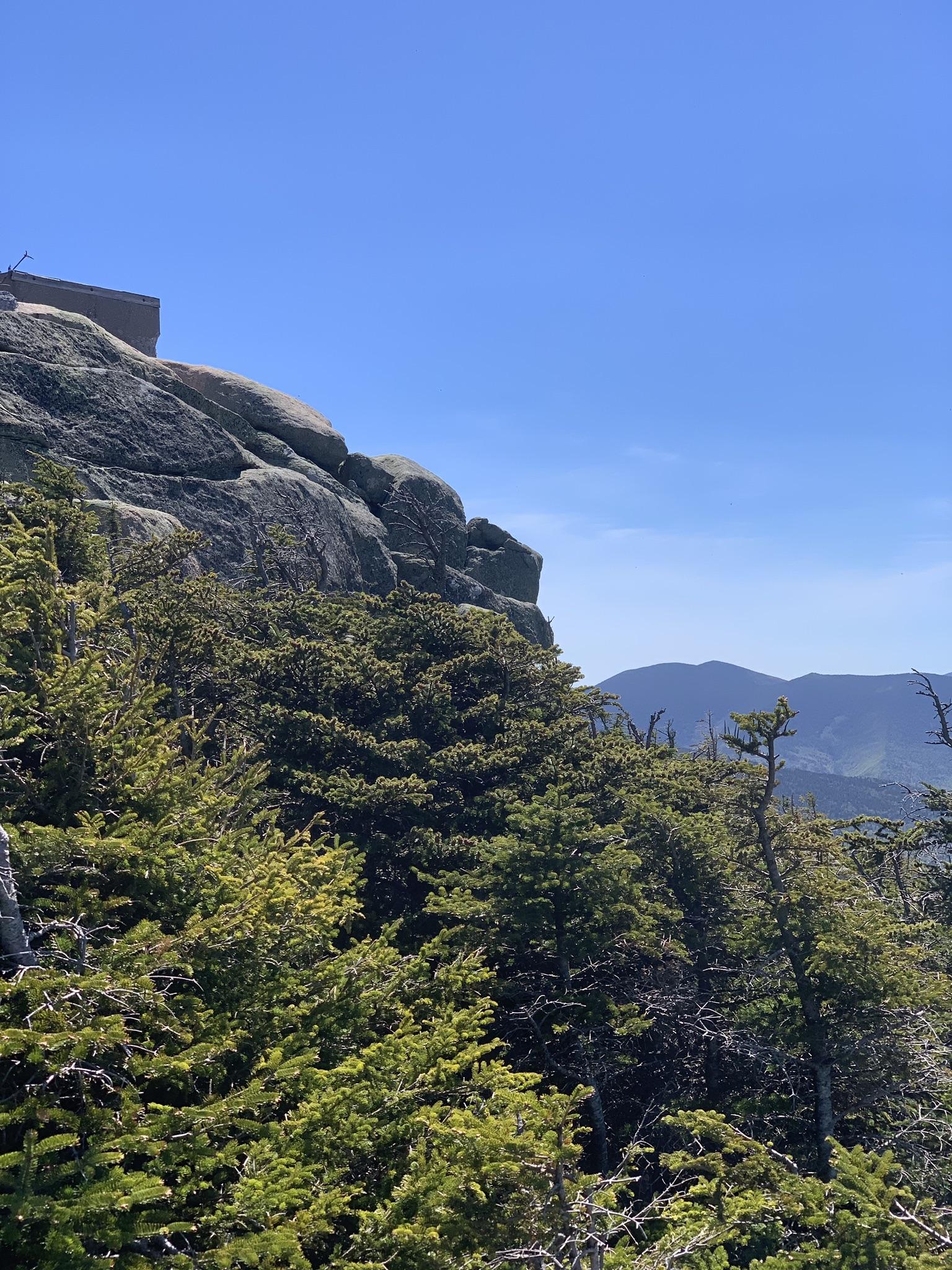 A building sits atop a rocky cliff overlooking evergreen trees and distant mountains.