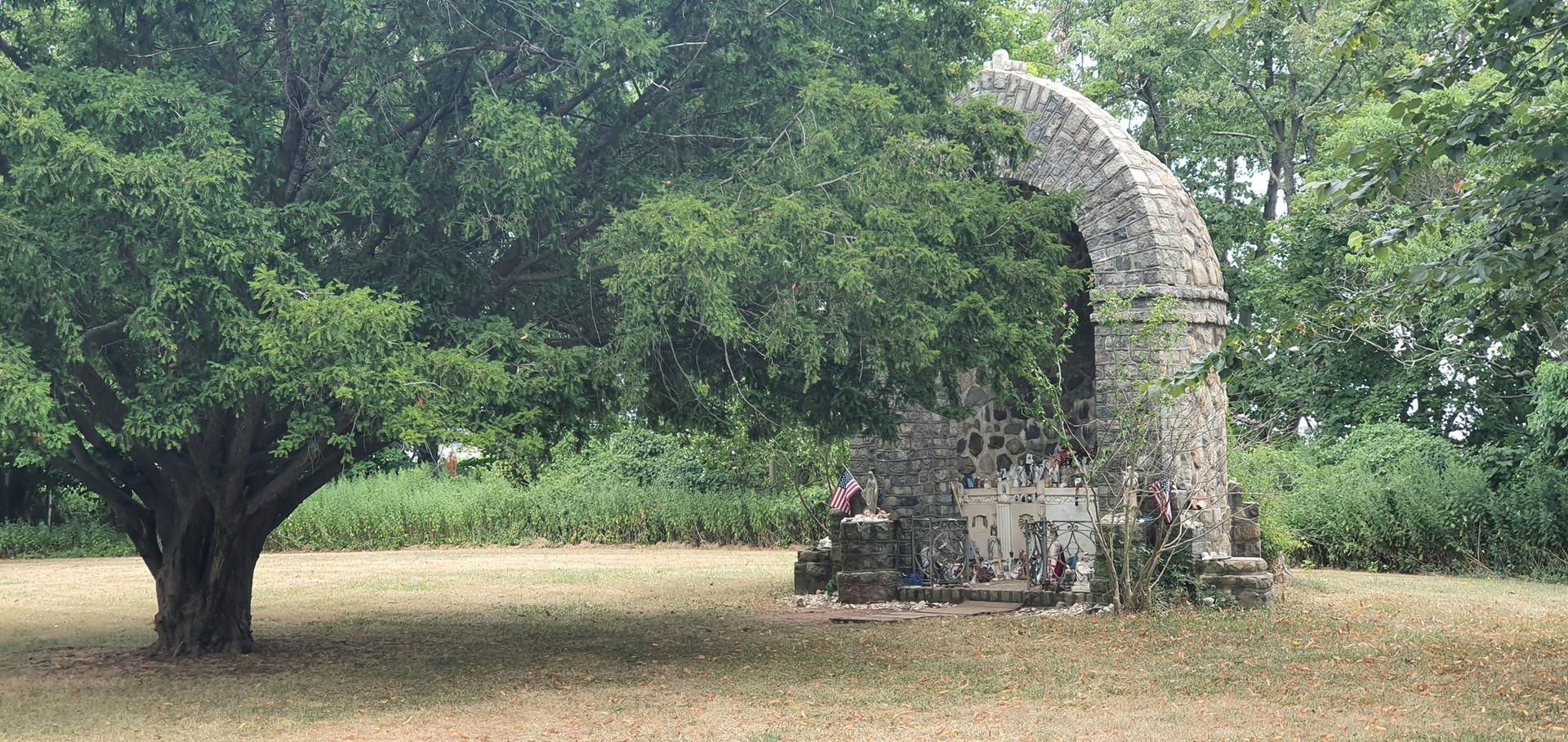Ancient stone archway ruin stands beside a large, gnarled tree in a field.