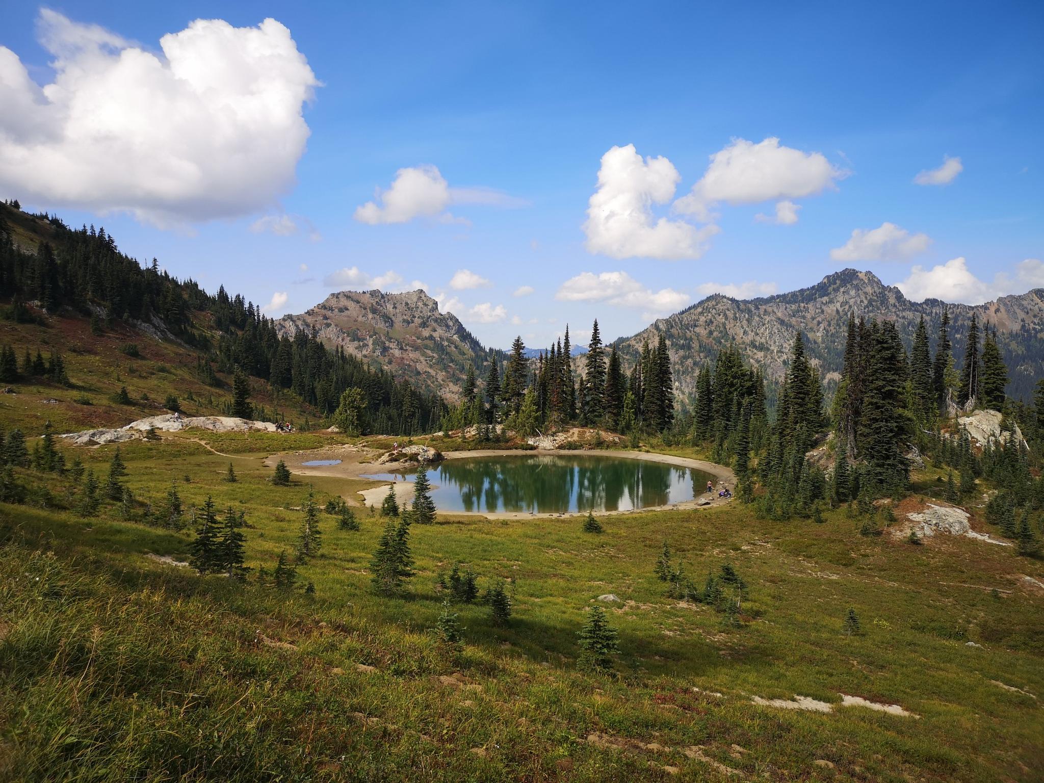 Small, calm lake reflecting a blue sky, surrounded by green meadows and mountains.