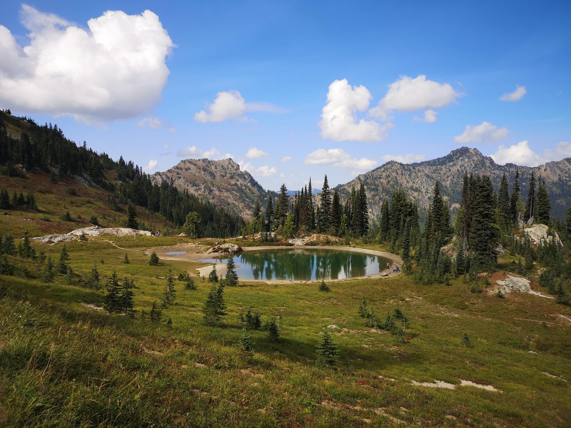 Small, calm lake reflecting a blue sky, surrounded by green meadows and mountains.