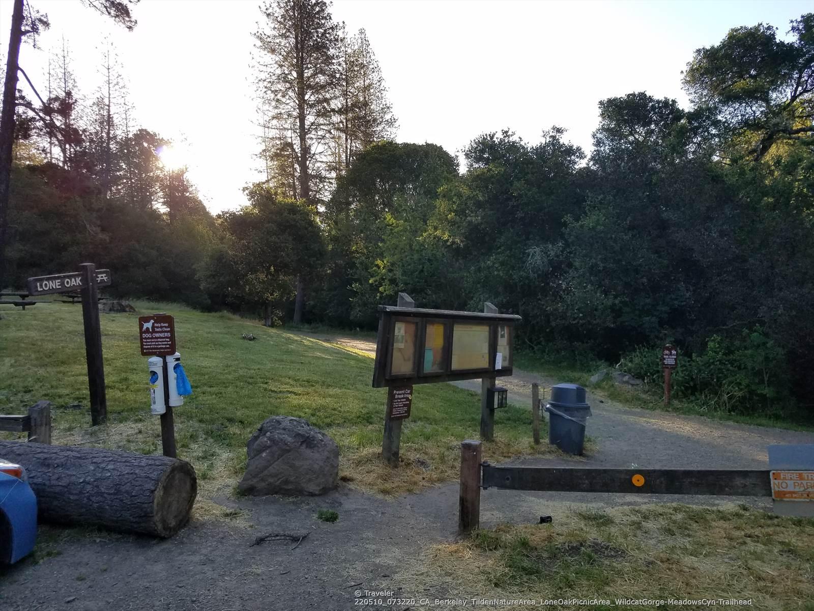 Trailhead with signs, trash can, and log, sun shining through trees.