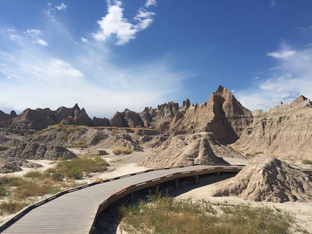 A wooden boardwalk winds through a sunlit, eroded badlands landscape under blue sky. - Fossil Exhibit Trail