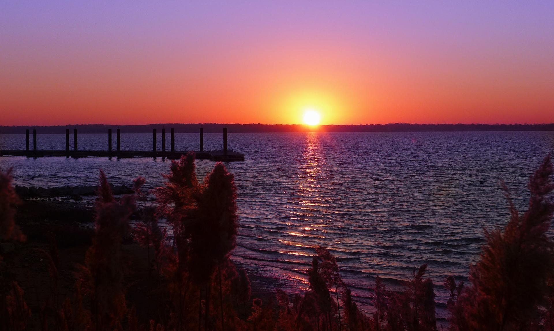 Vibrant sunset over water with a dock and foreground plants.
