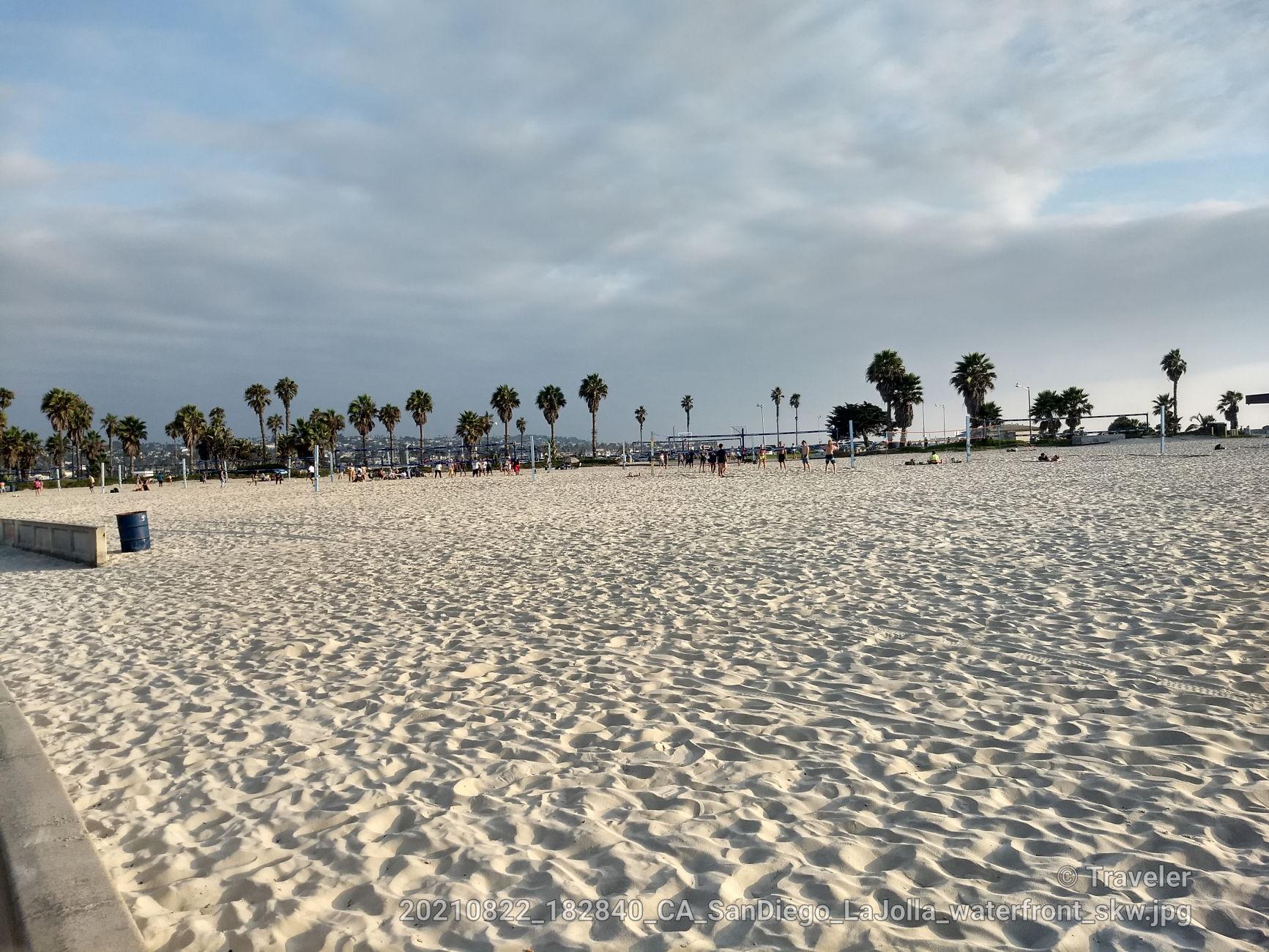 Expansive sandy beach with distant palm trees and people playing volleyball.