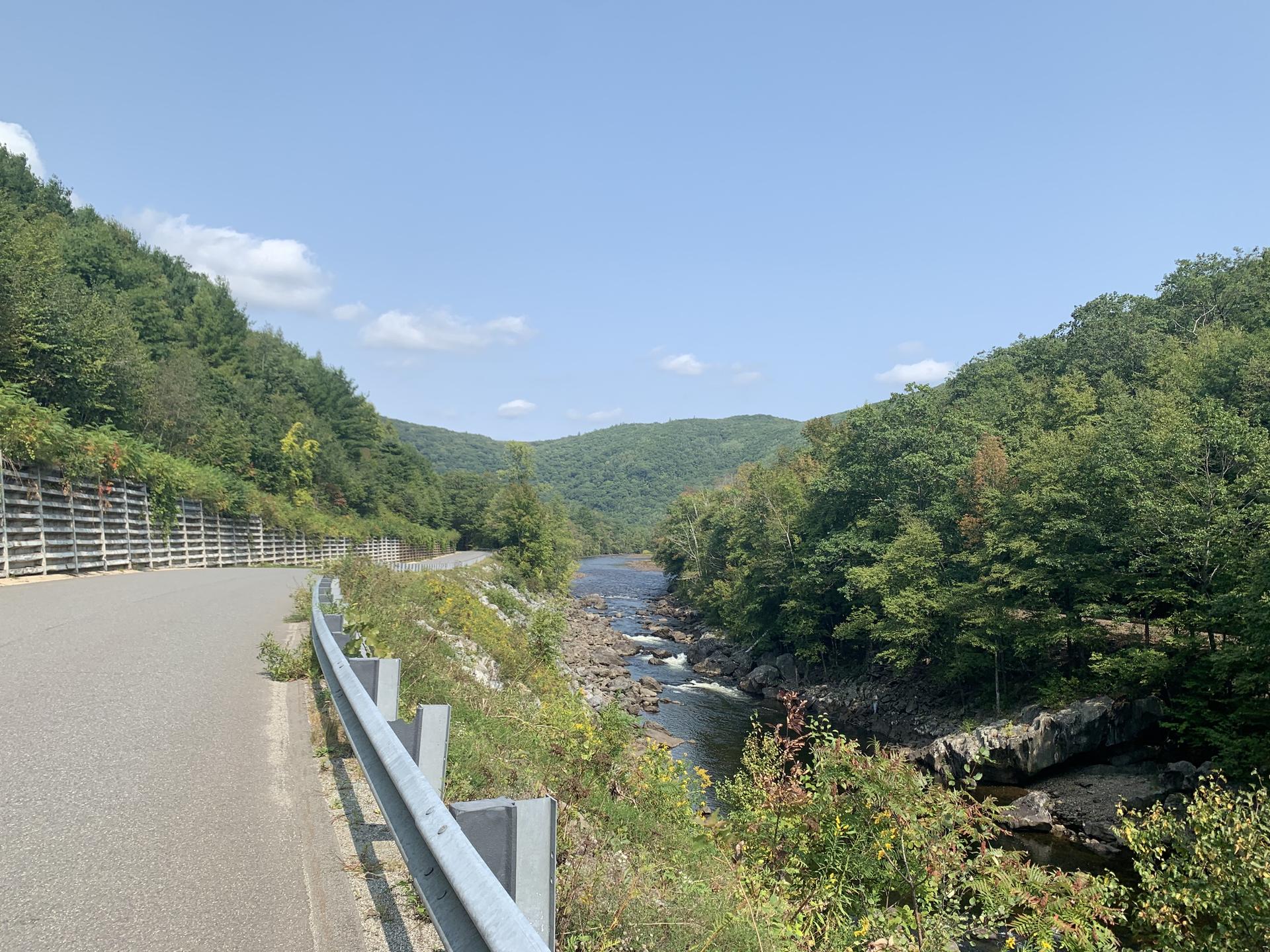 A winding road alongside a river in a forested valley on a sunny day.