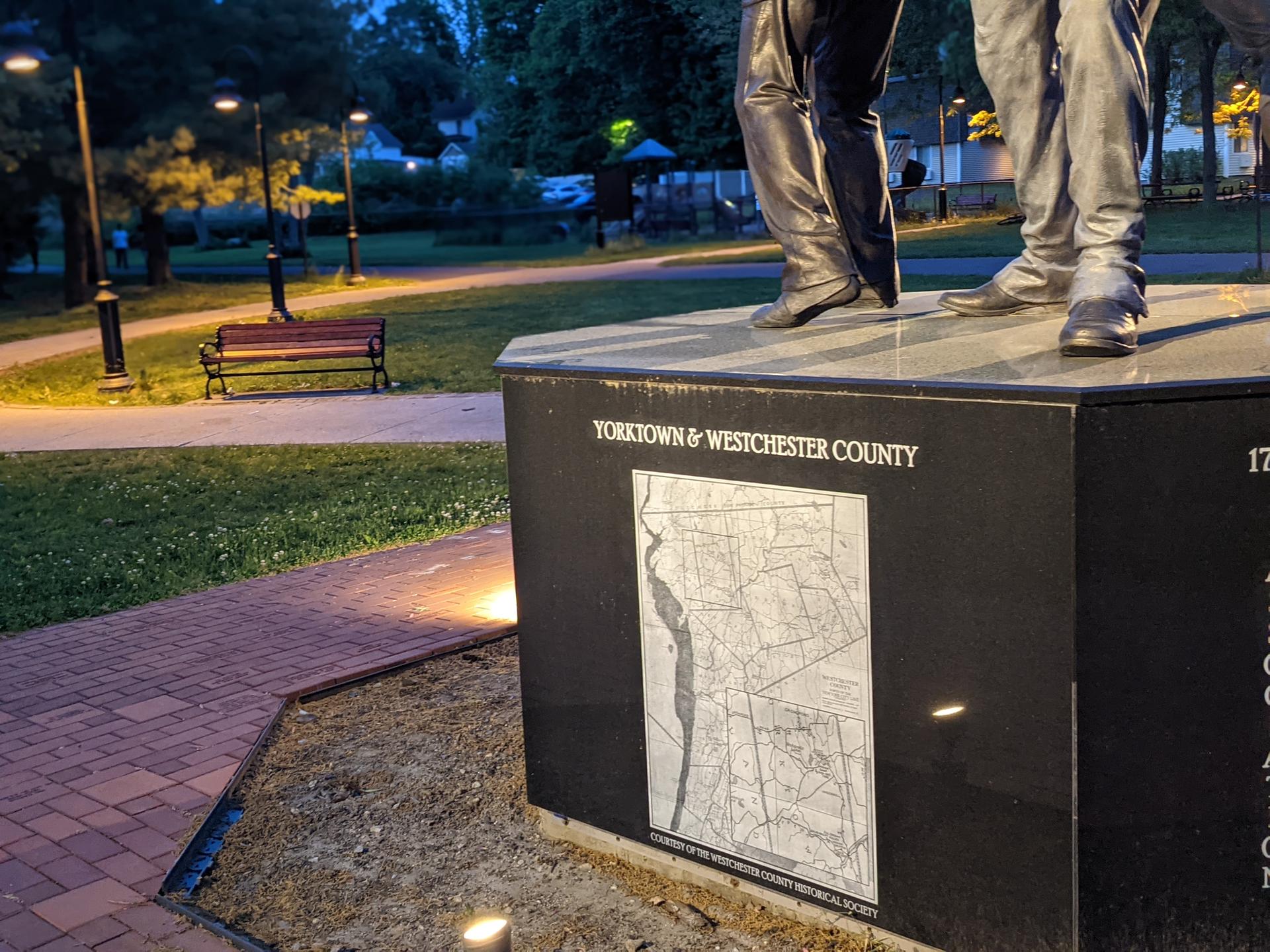 Statue base with map in a park at dusk, streetlights glowing.