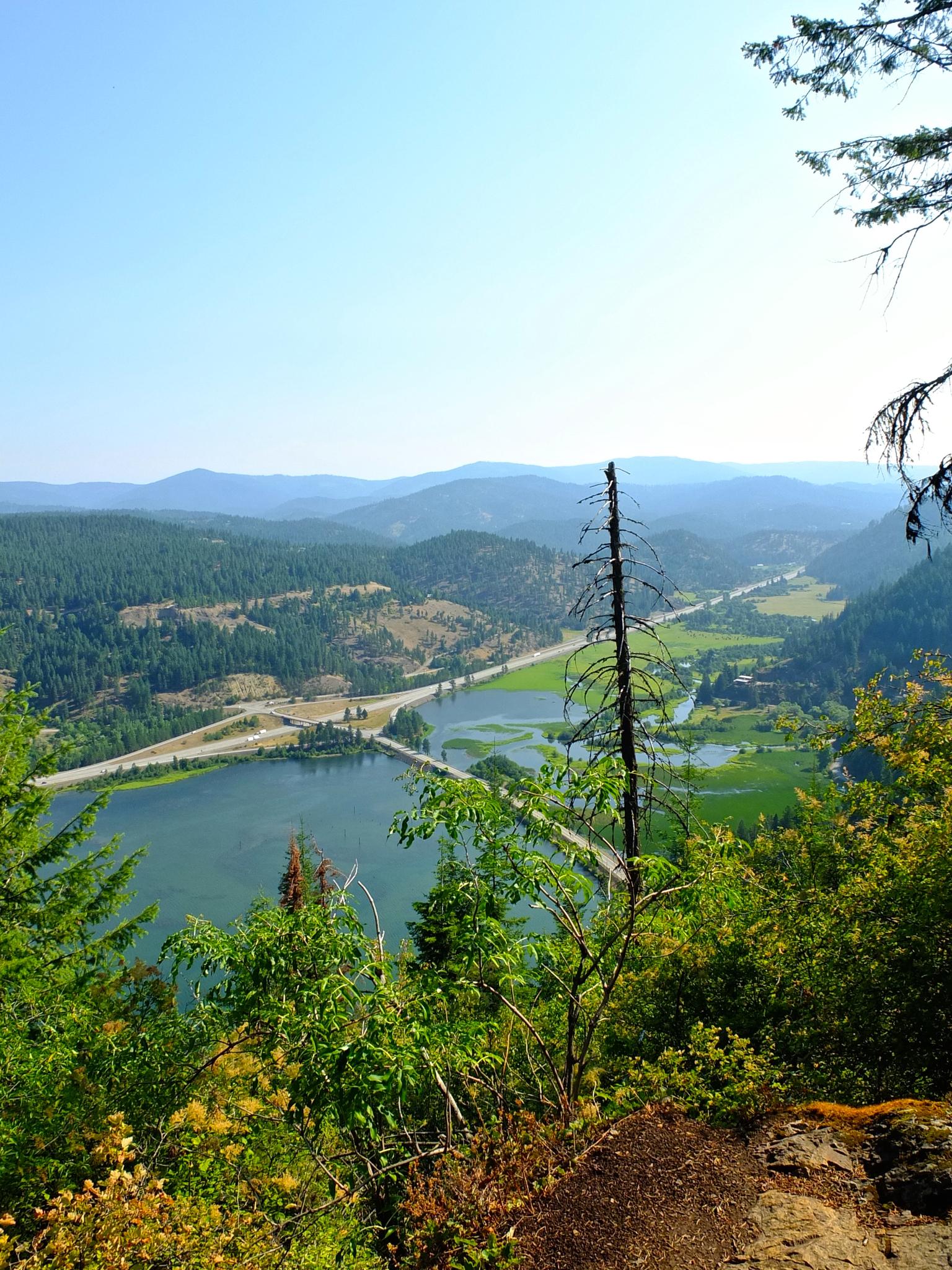 Bright valley view of a lake, highway, and forested mountains under clear sky.