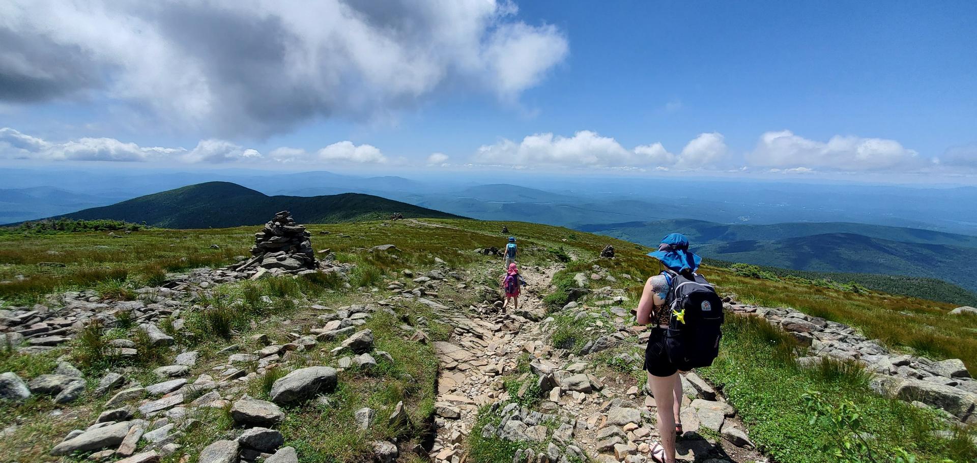 Hikers on a rocky mountain path with a cairn and expansive views.
