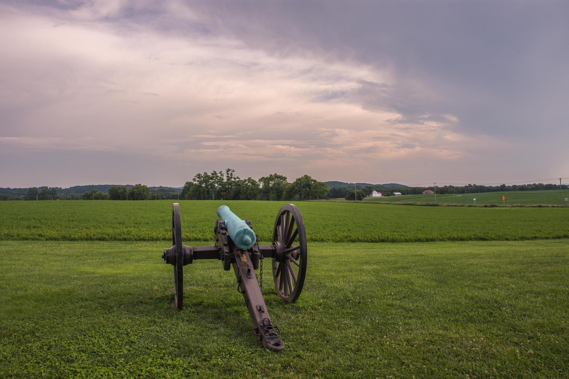 An antique cannon stands in a vast green field under a cloudy sky.