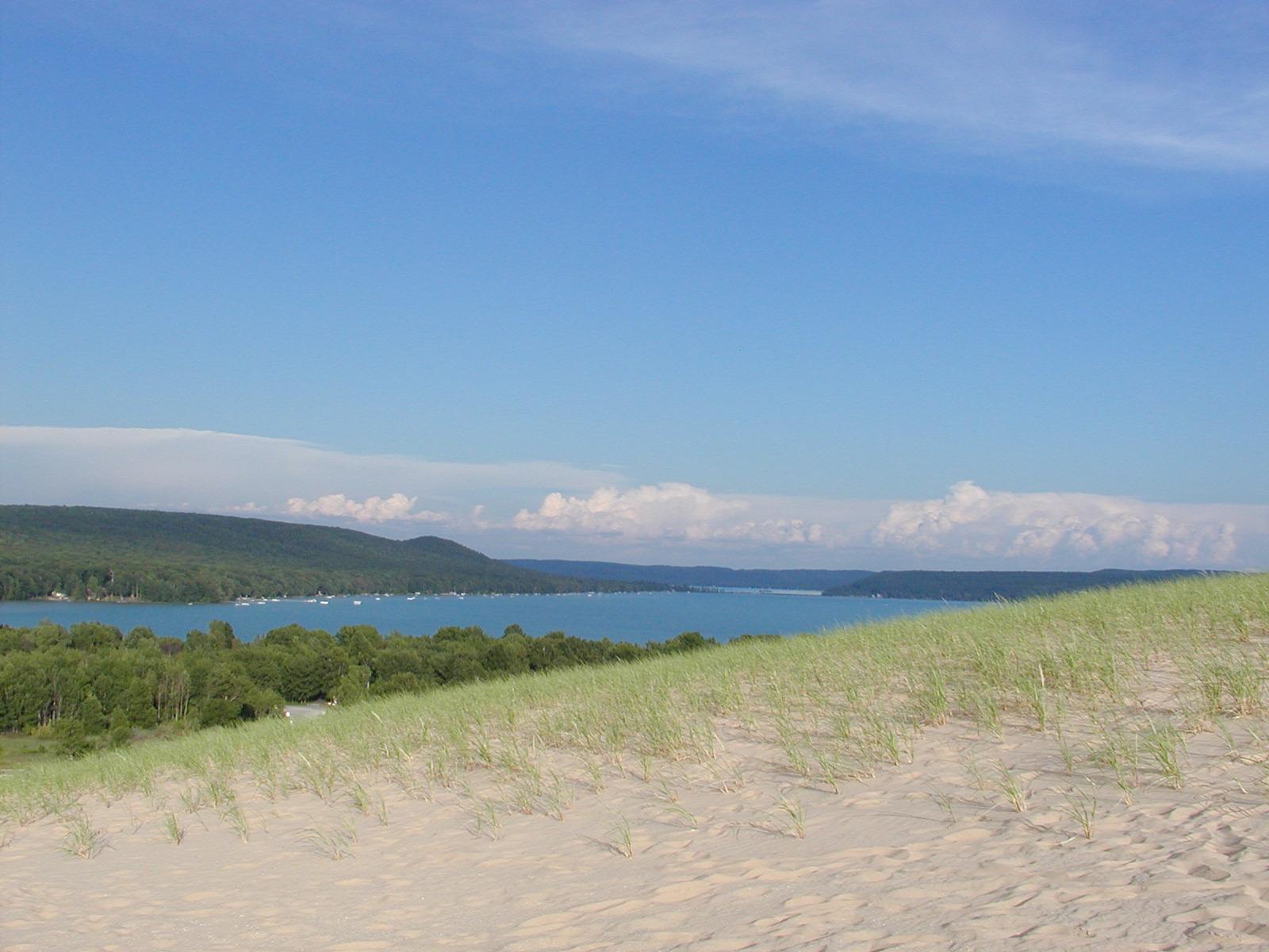 Grassy sand dune overlooks a large lake with boats and forested hills.