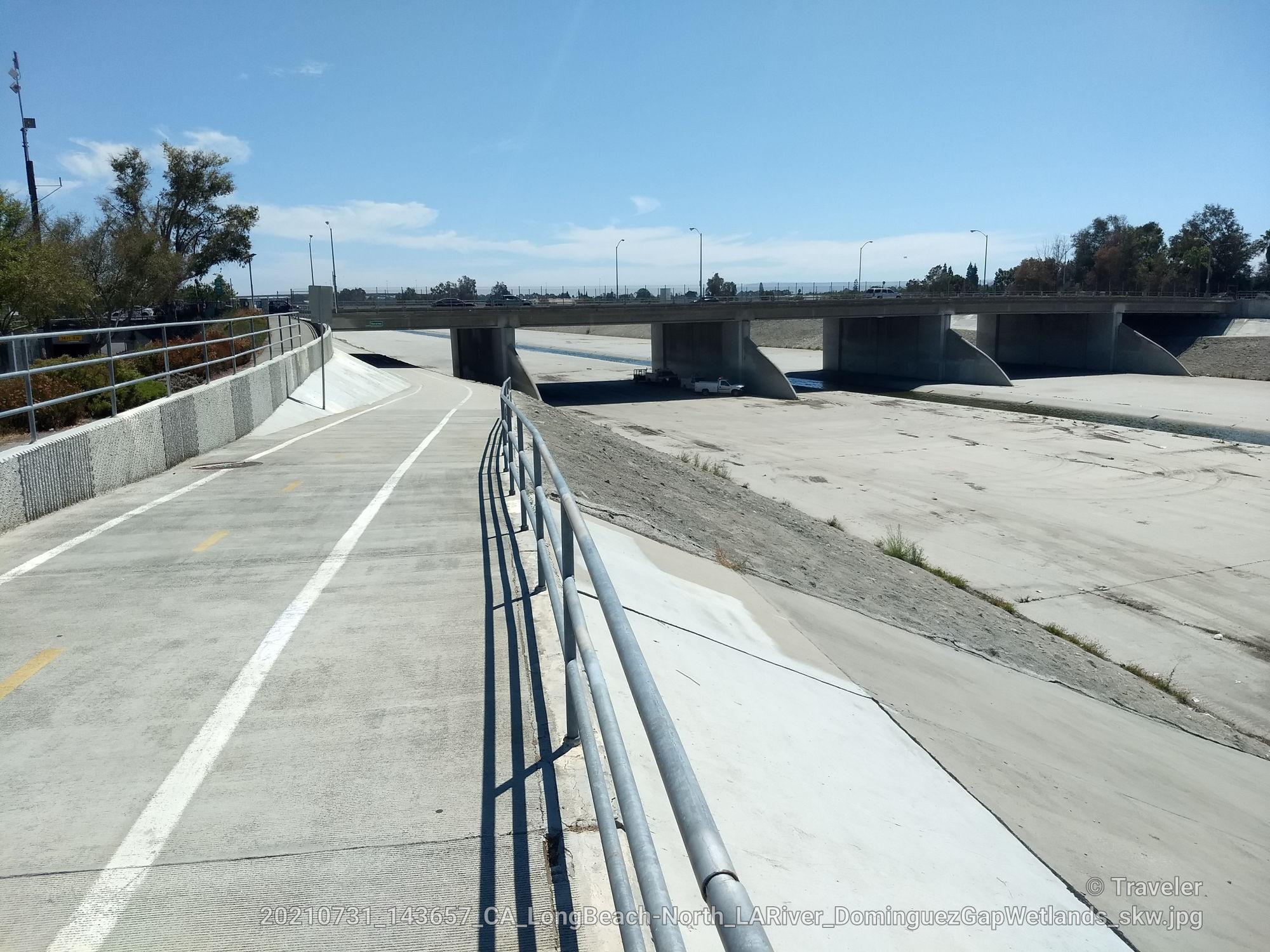 Paved path alongside a dry concrete channel leading to a large bridge.
