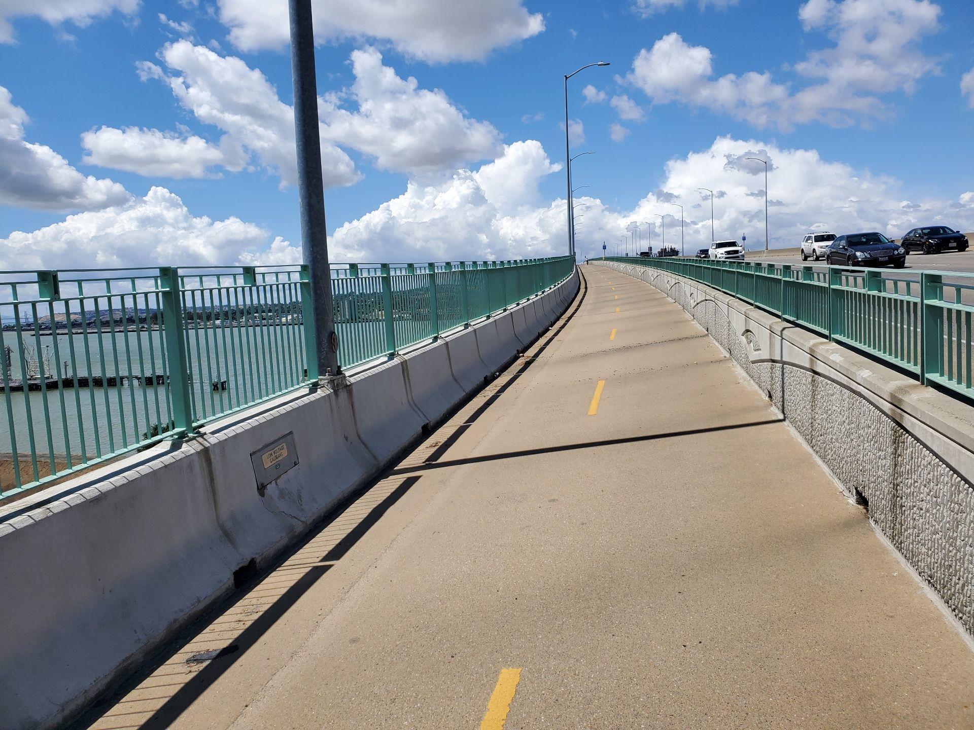 Empty paved path on a bridge with green railings under a blue sky.