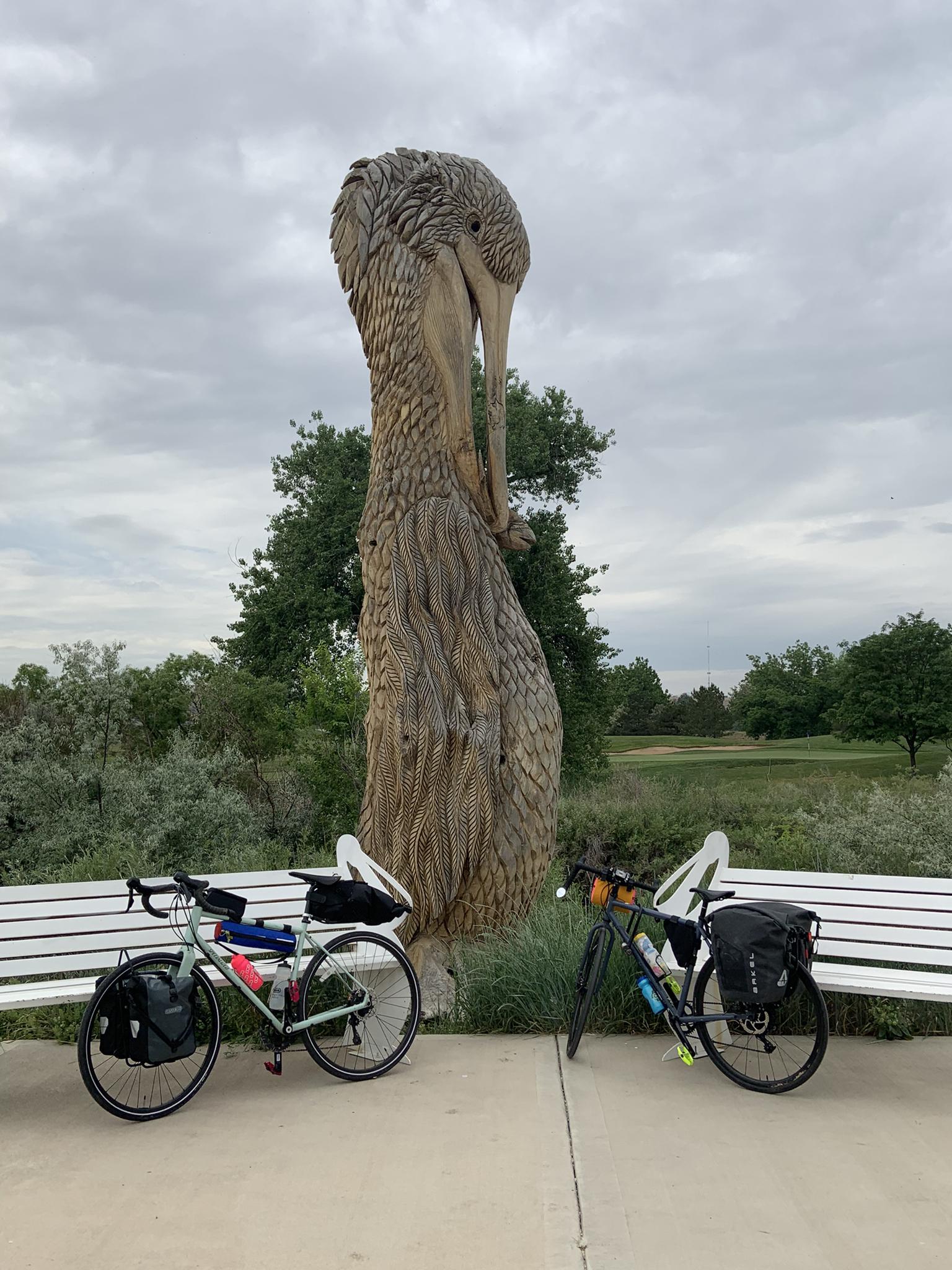 Two bicycles parked next to a tall wooden bird sculpture on an overcast day.