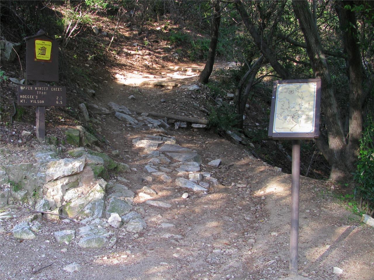 A rocky dirt trail with two wooden signs in a sun-dappled wooded area.
