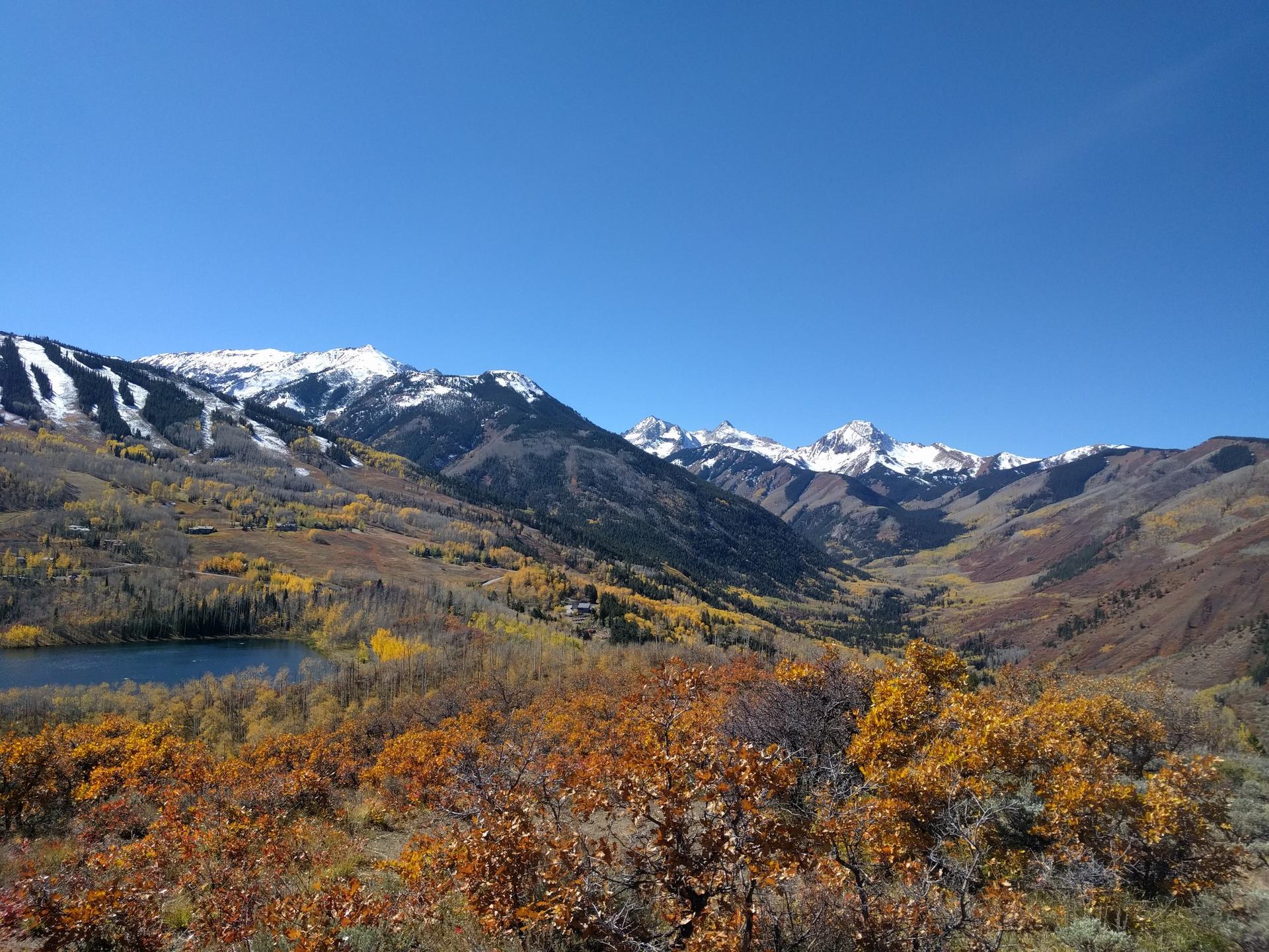 Snow-capped mountains, a lake, and vibrant autumn trees under a clear sky.