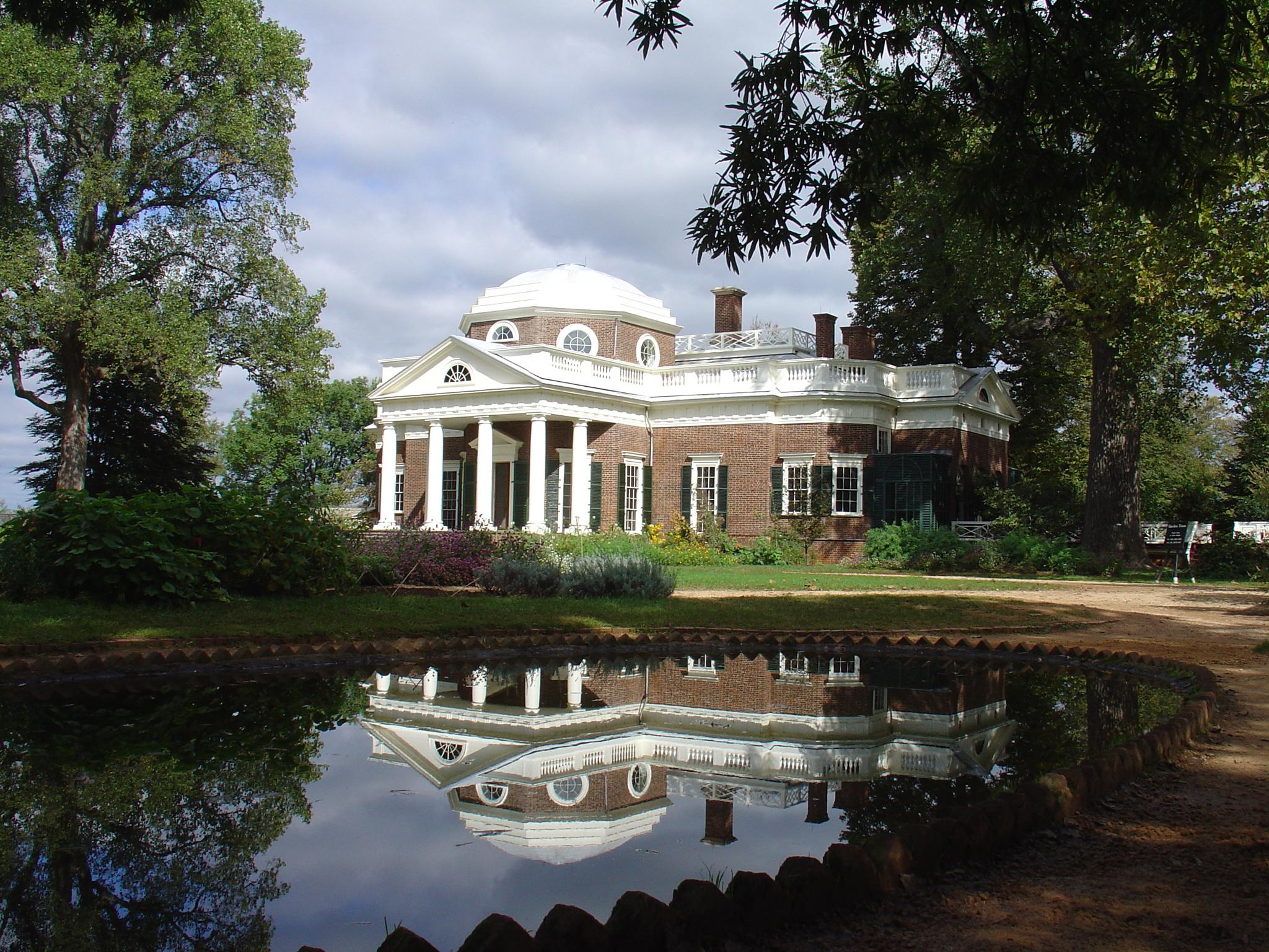 Classical brick building with white dome reflected in a pond under a cloudy sky.
