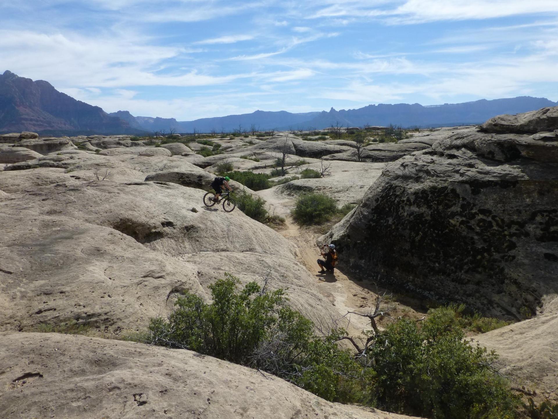 Two mountain bikers on rocky desert terrain with large formations and distant mountains.