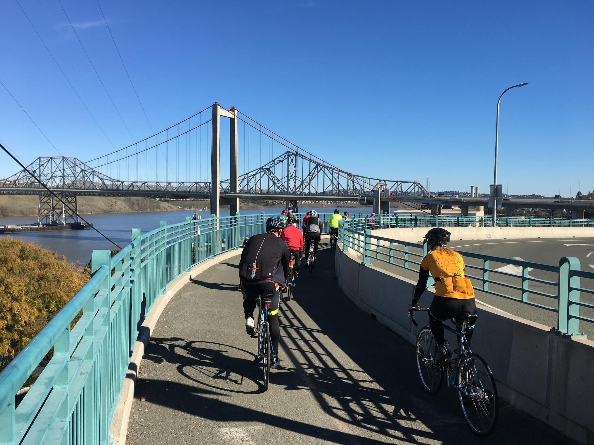 Cyclists ride on a paved path towards a large suspension bridge over water. Bright day.