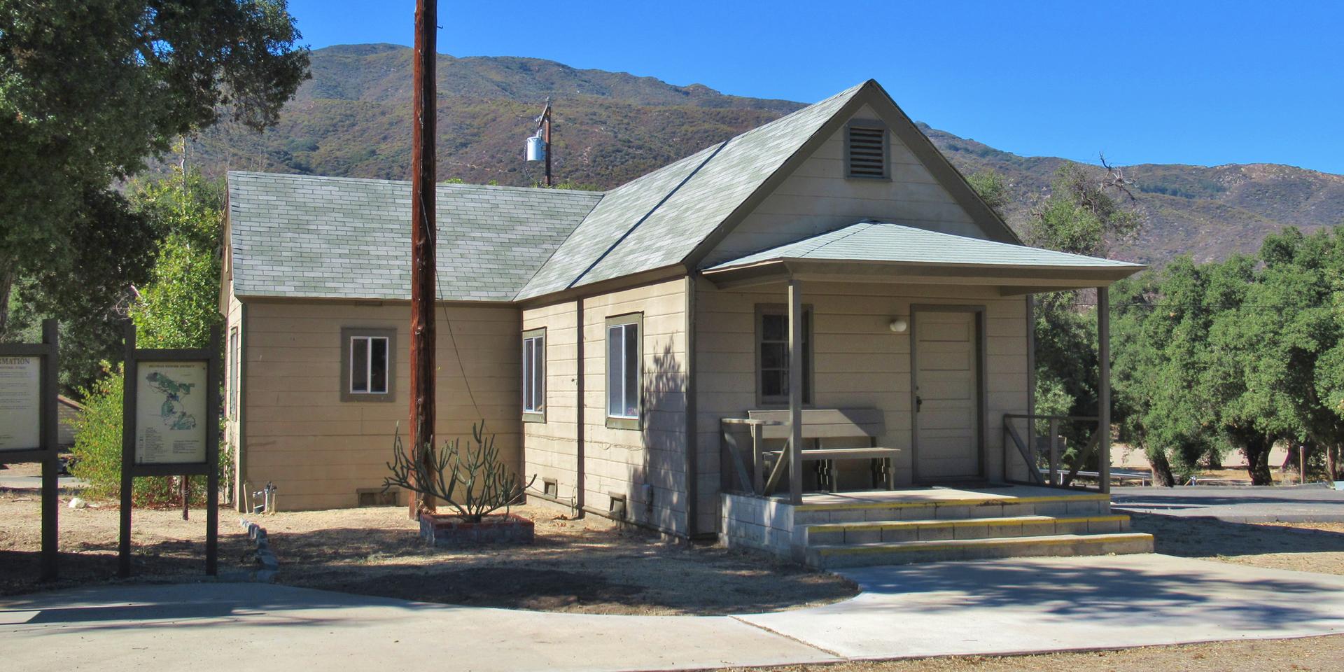 Small, light-colored house with a porch, mountains in background, under clear sky.