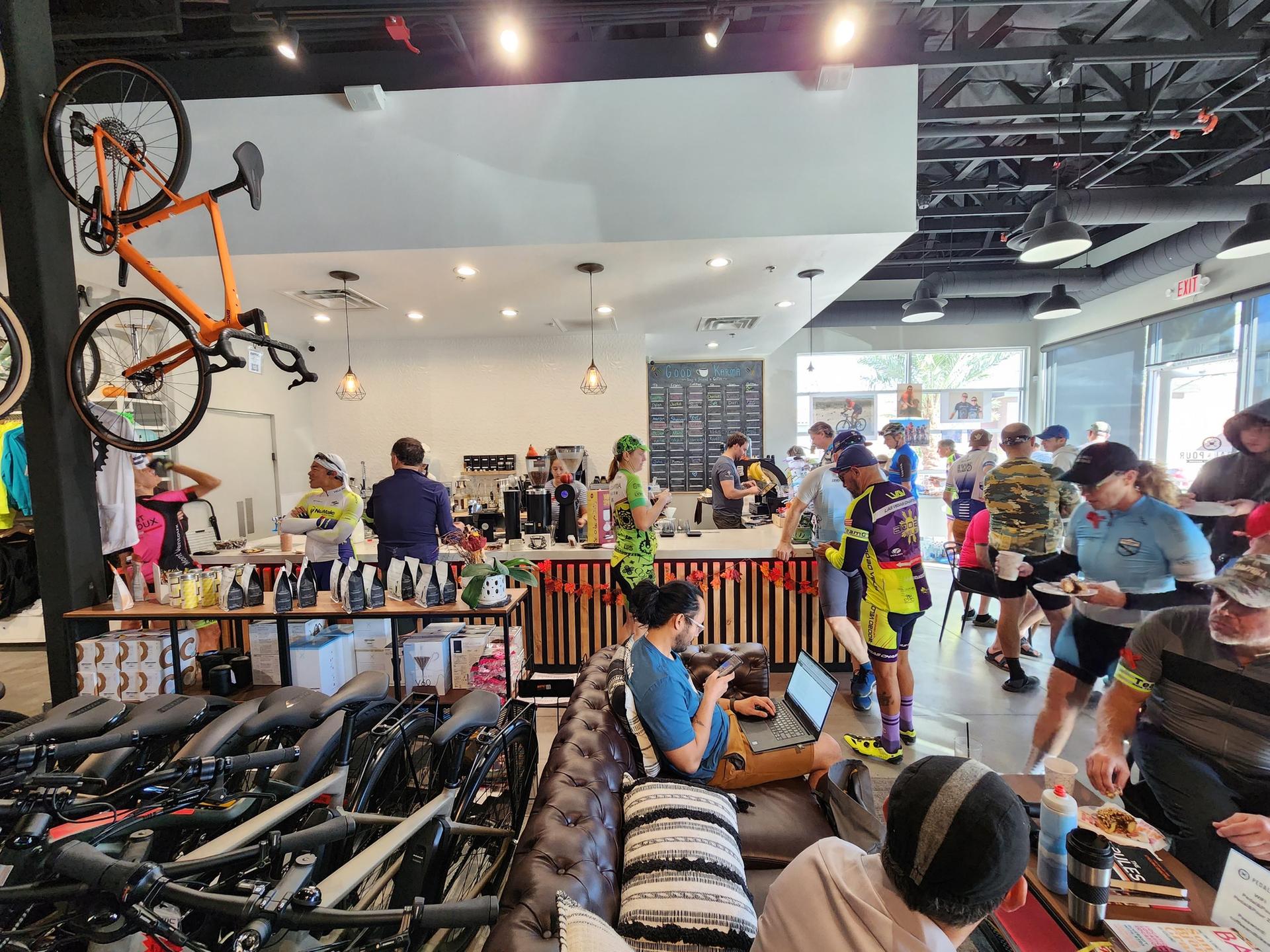 A busy cafe and bike shop with many people socializing and an orange bicycle hanging.