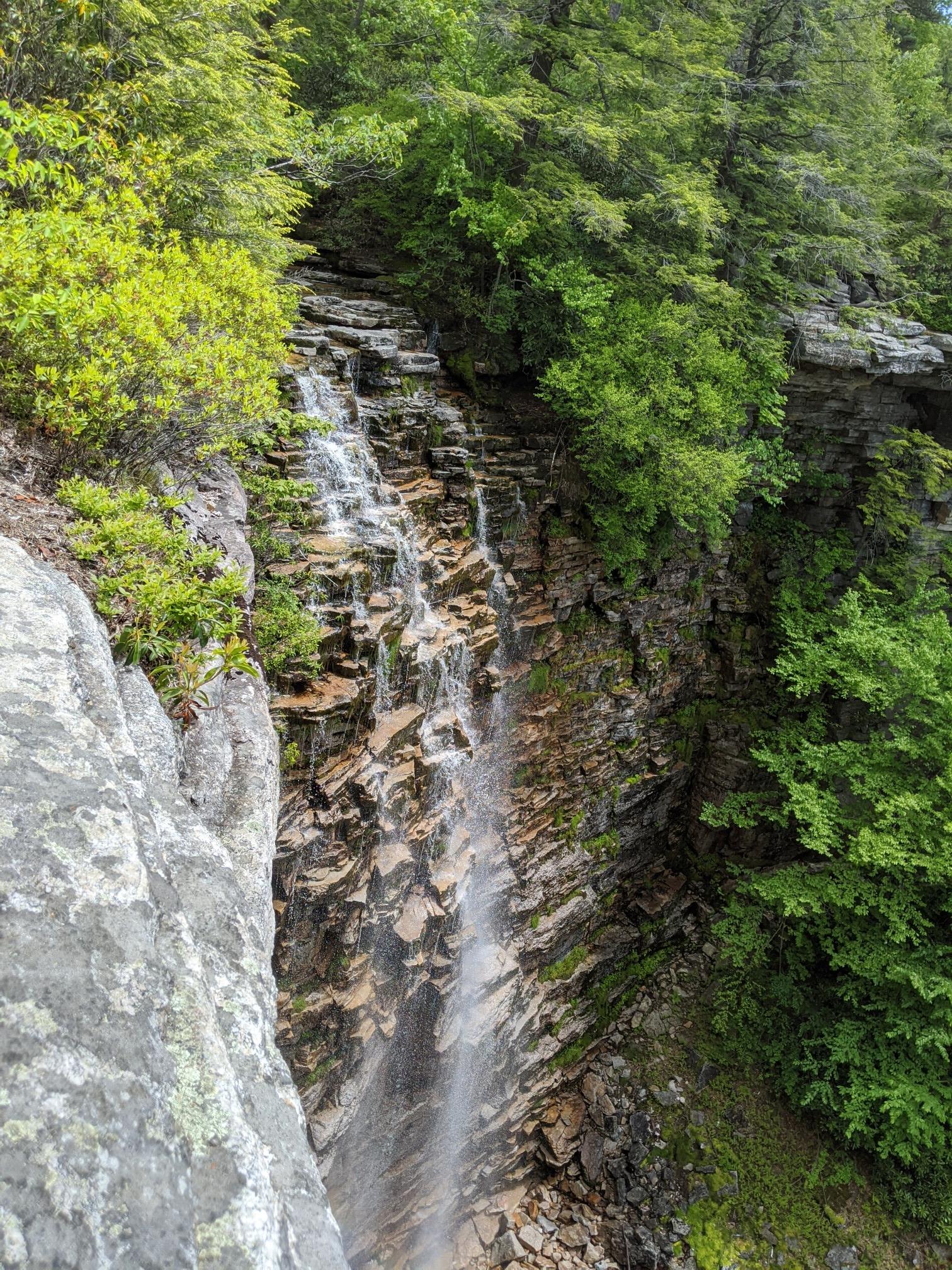 A waterfall cascades down a layered rock cliff surrounded by green foliage.