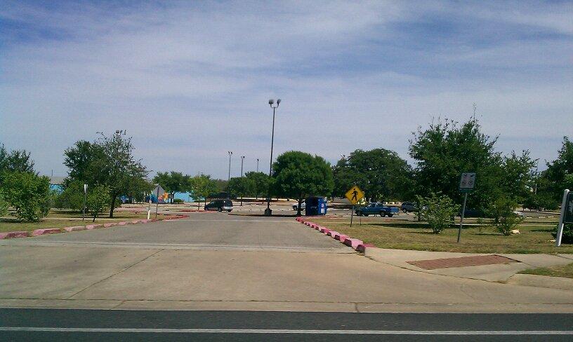 Wide asphalt entrance to a parking lot with trees under a bright sky.