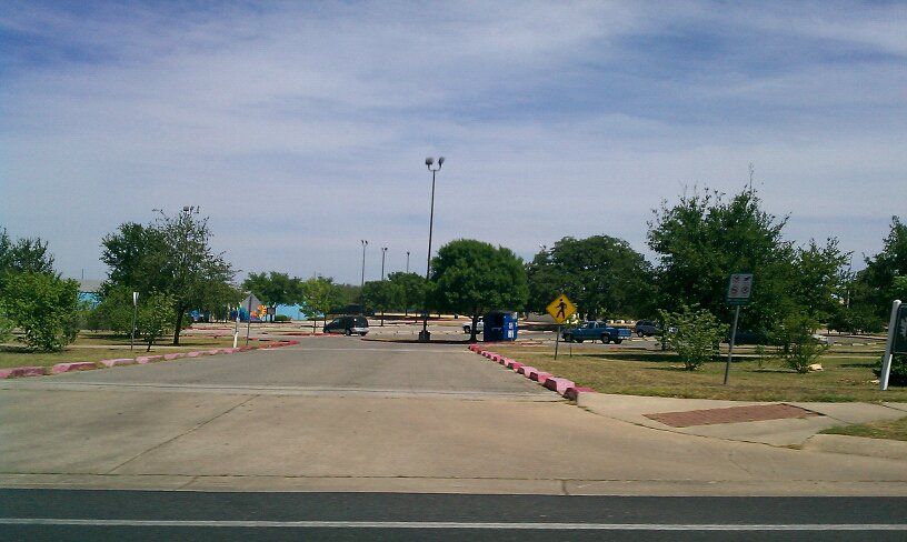 Wide asphalt entrance to a parking lot with trees under a bright sky. - Dick Nichols District Park loop from NXP Oak Hill Fab