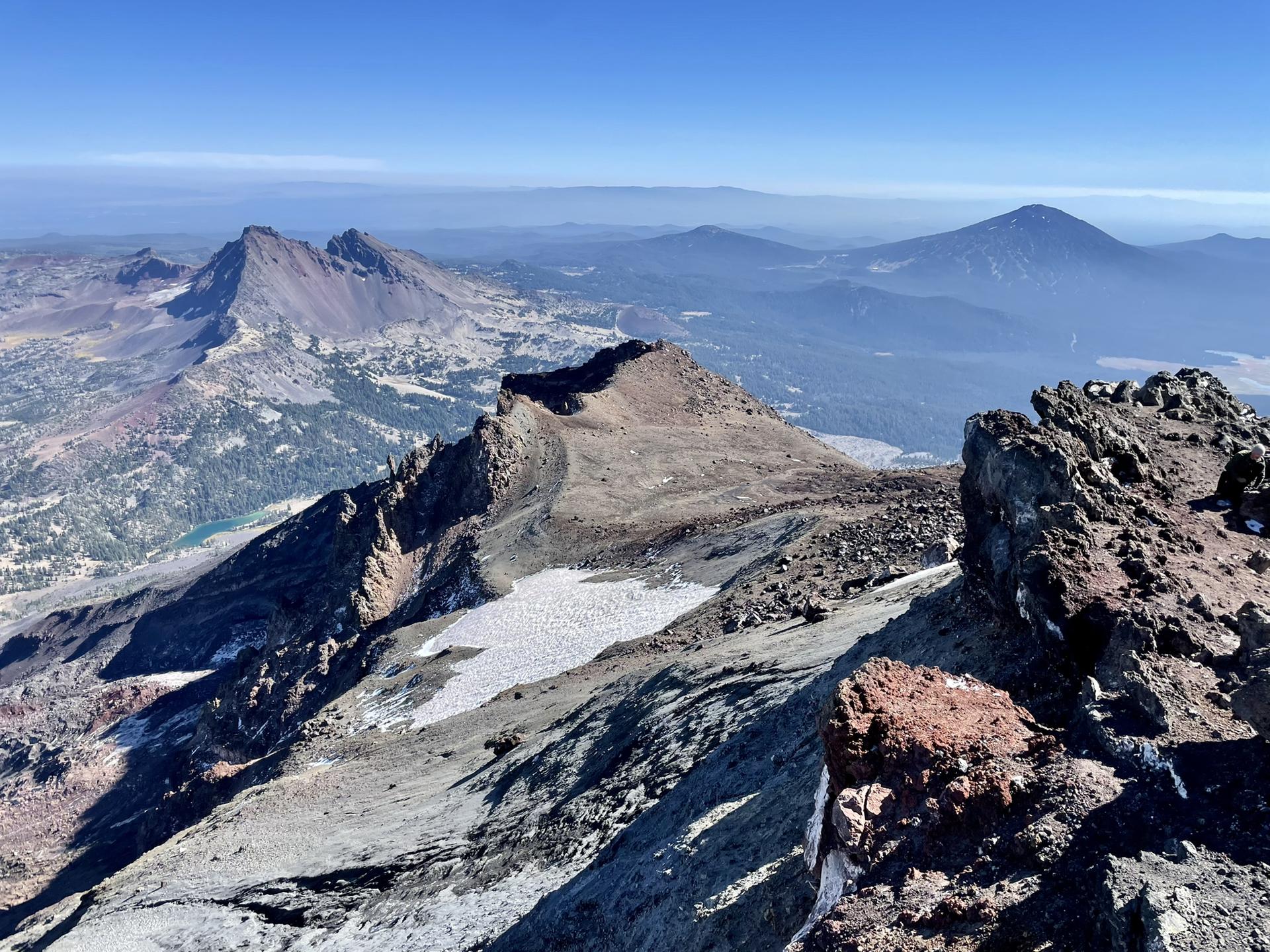 Panoramic view from a rocky mountain peak with snow patches and distant peaks.