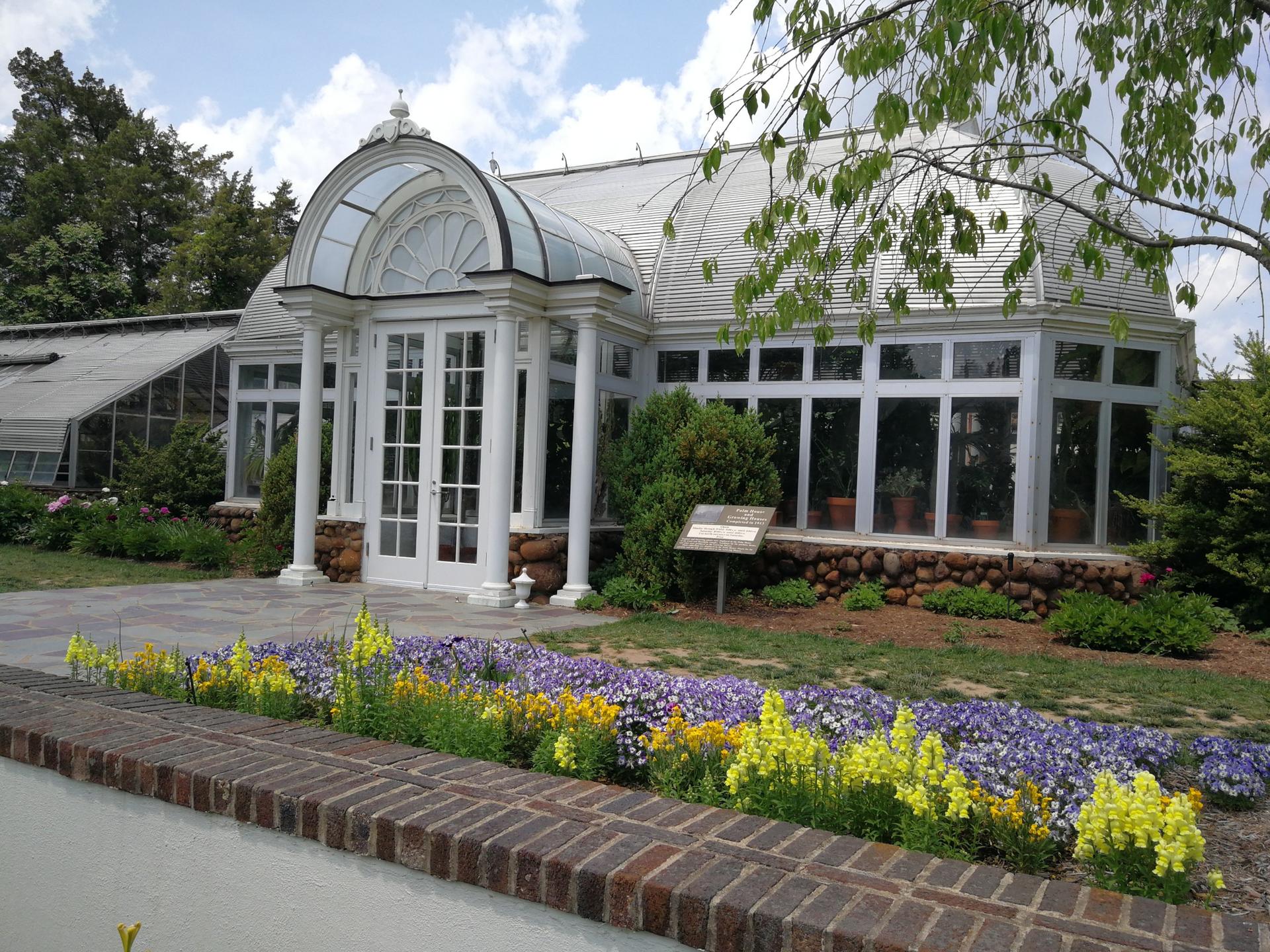 Elegant glass greenhouse with white trim, surrounded by colorful flower beds under bright daylight.