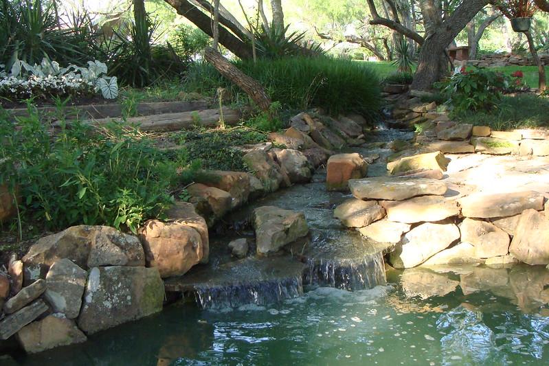 A small waterfall cascades over rocks into a pond, surrounded by lush greenery.