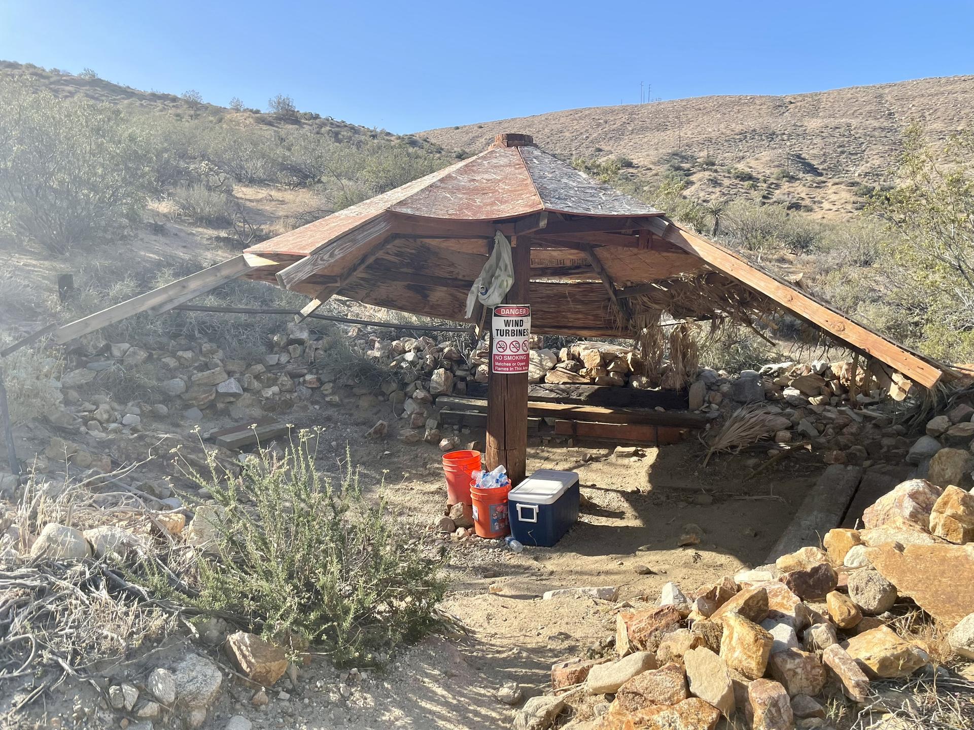 Rustic wooden shelter with buckets and cooler in a sunny, arid landscape.