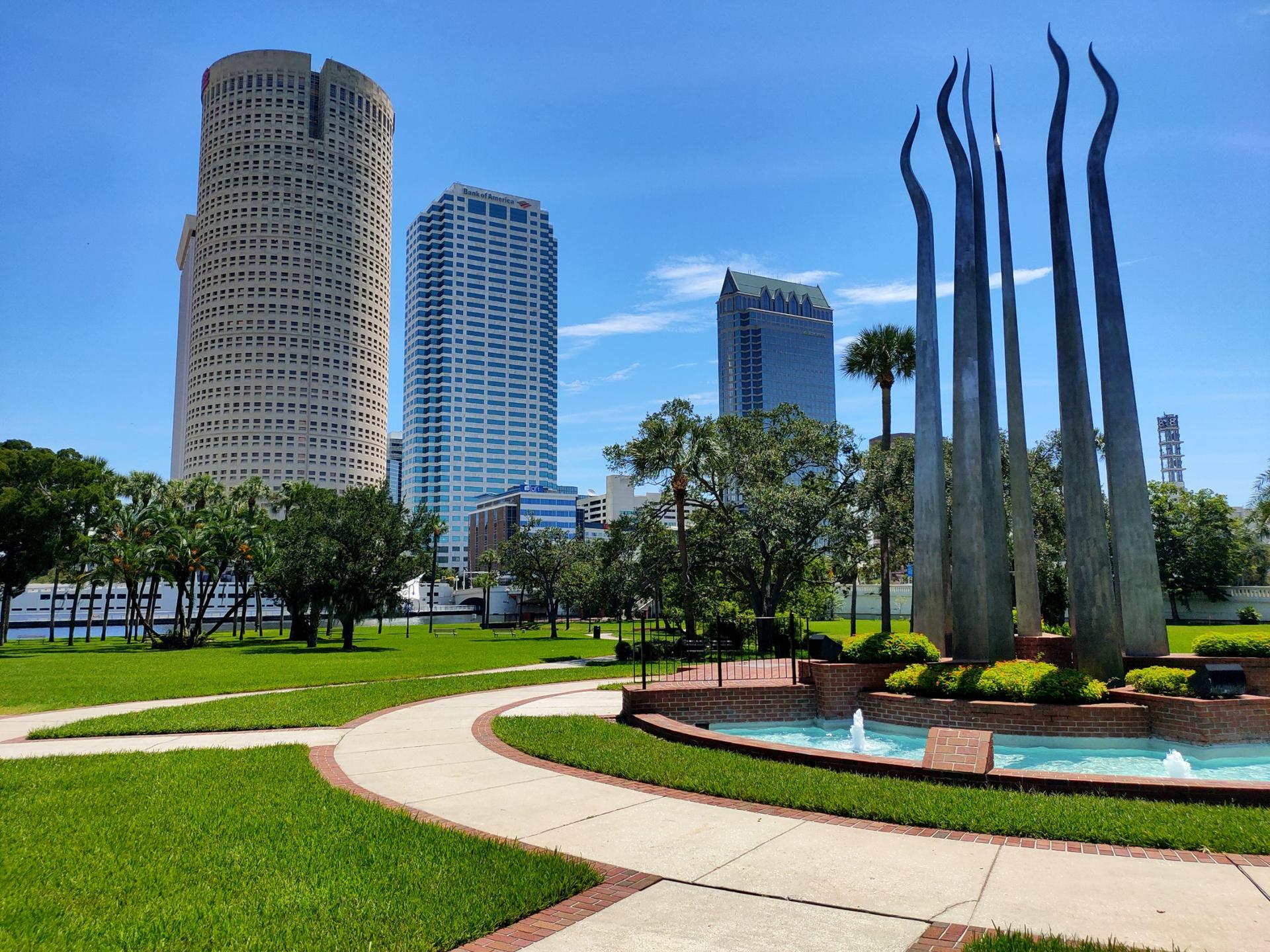 Modern city buildings overlook a park with a tall sculpture and fountain.