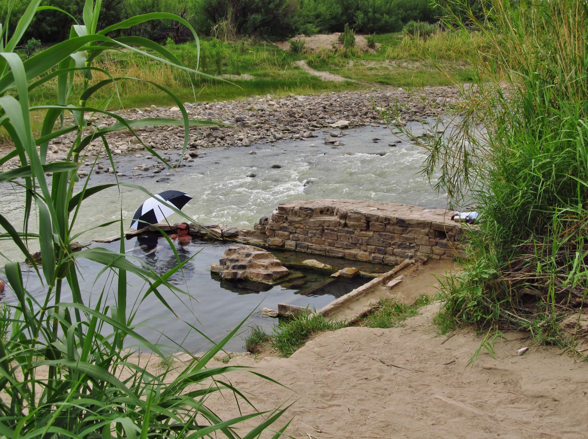 People relax in a river next to a stone structure, surrounded by green reeds.