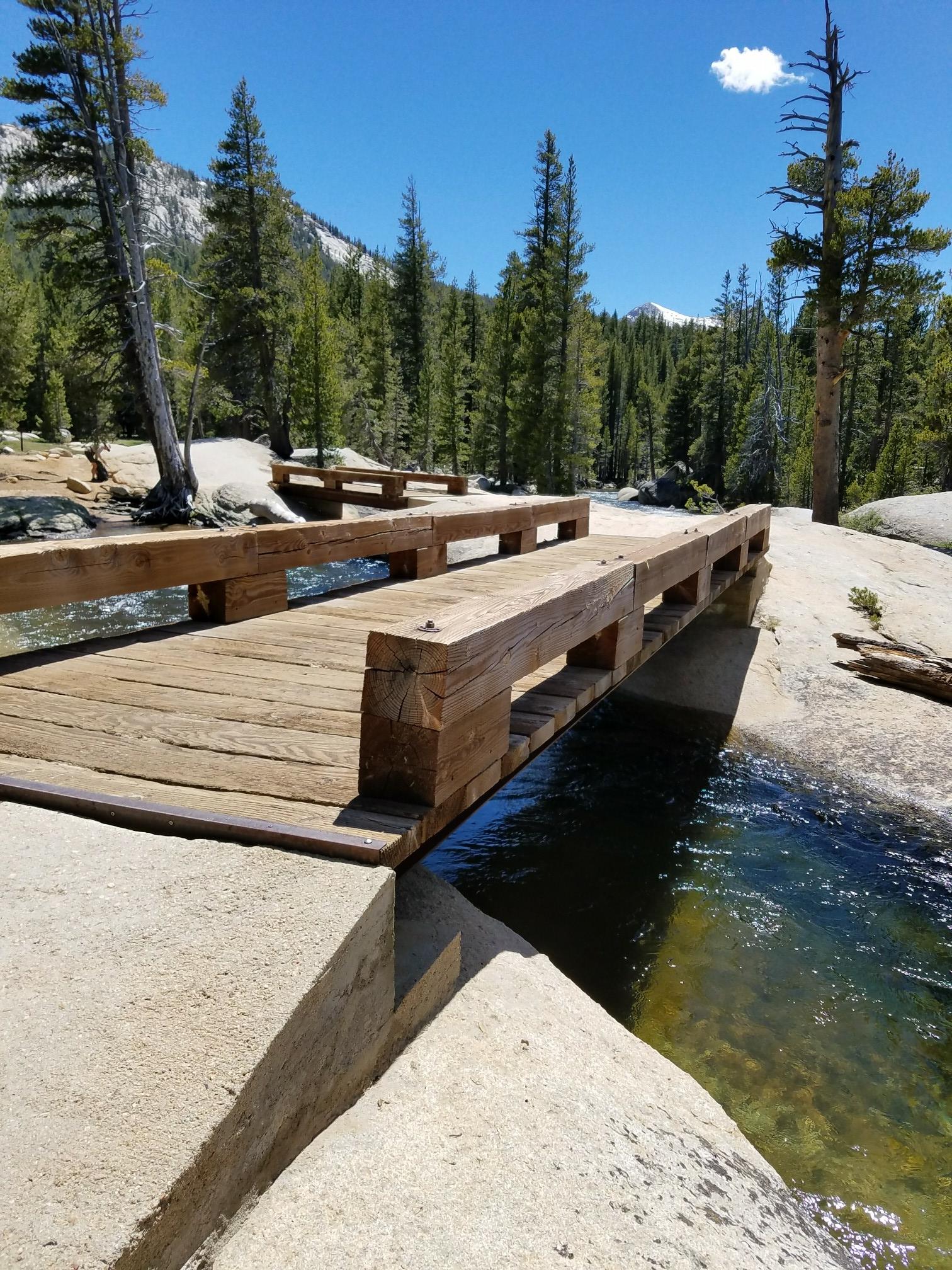 Wooden bridge crosses a clear stream, surrounded by granite rocks and pine trees.