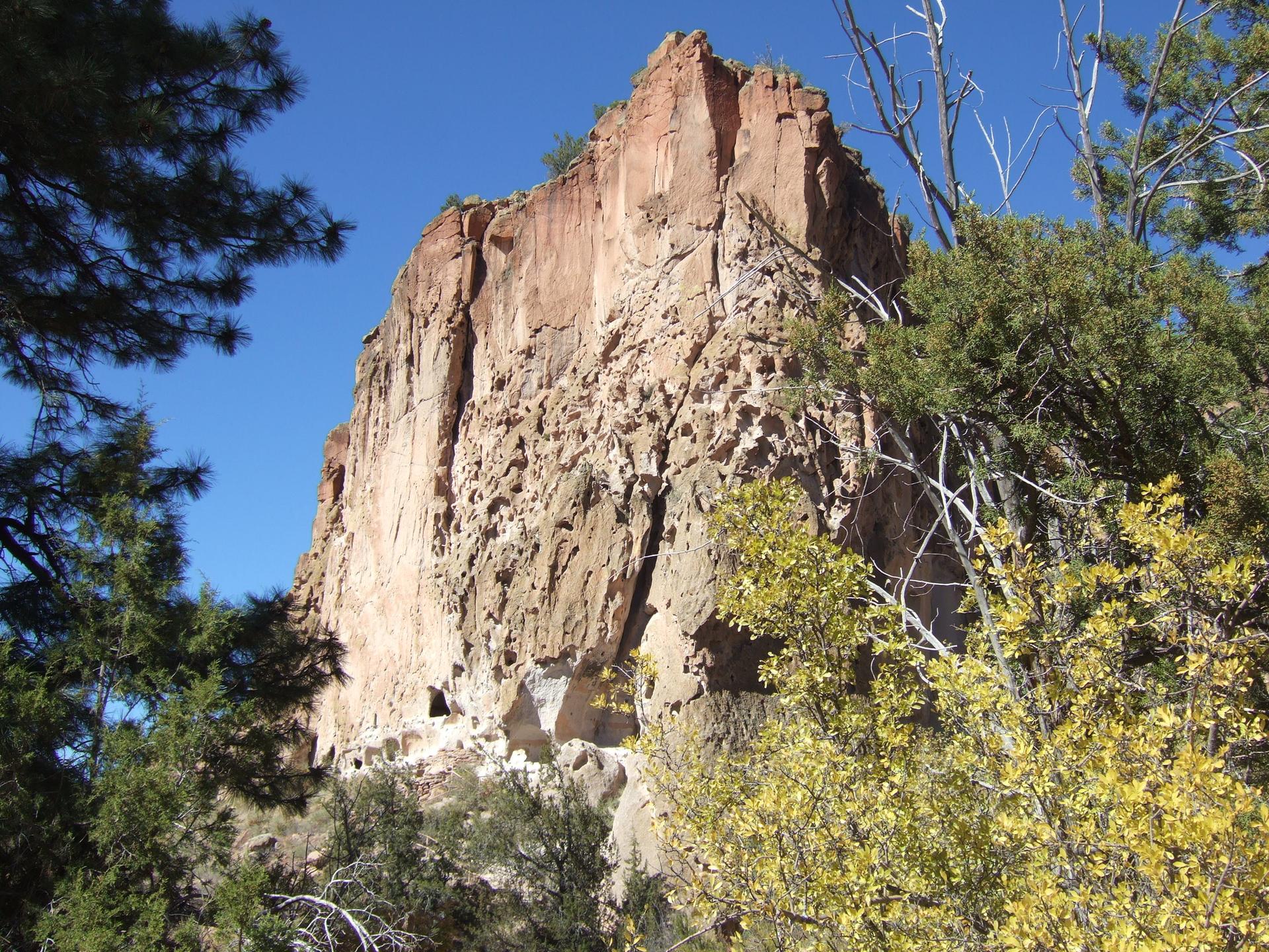 A massive, sunlit reddish-brown rock formation towers over green and yellow trees.