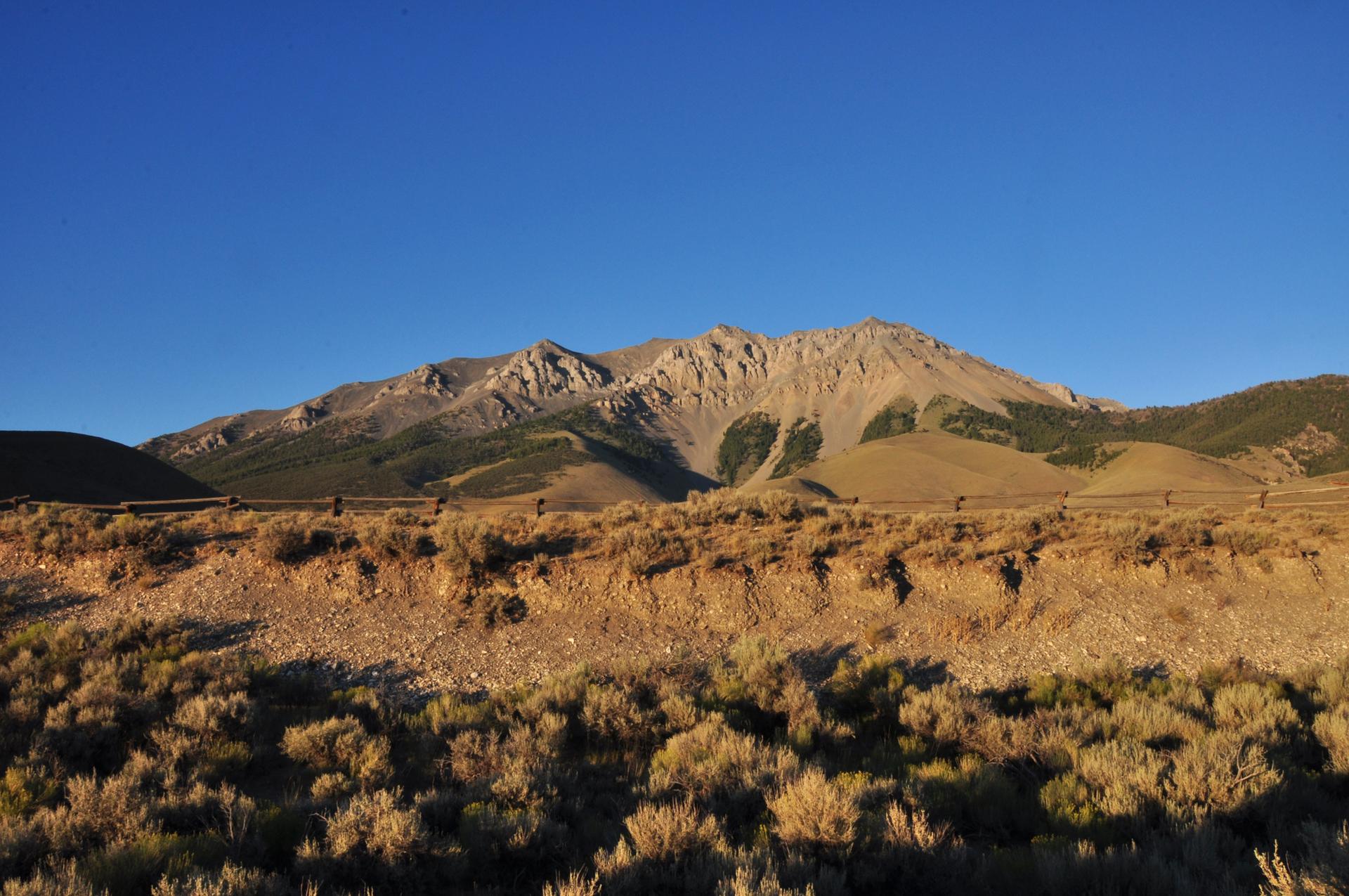 Sunlit rocky mountains overlook dry, brushy hills under a bright blue sky.