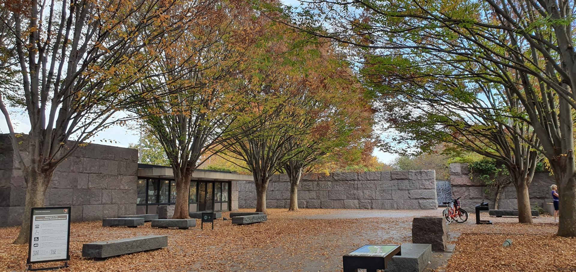 An outdoor plaza with autumn trees, stone walls, and a person on a bike.