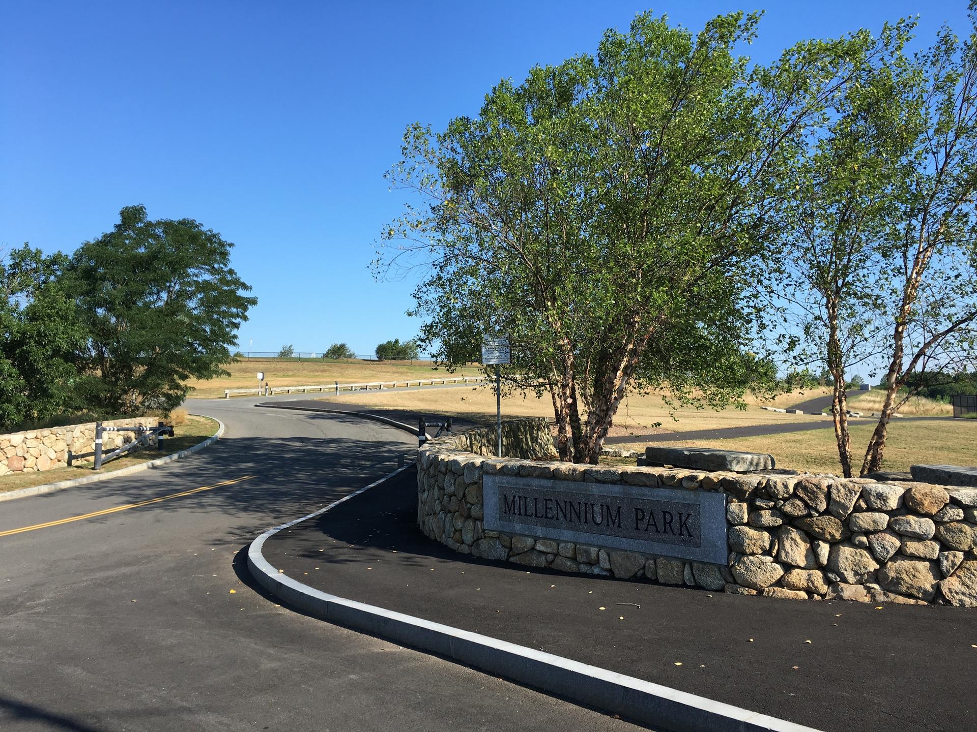 A paved road curves past a stone wall and green trees under a bright blue sky.