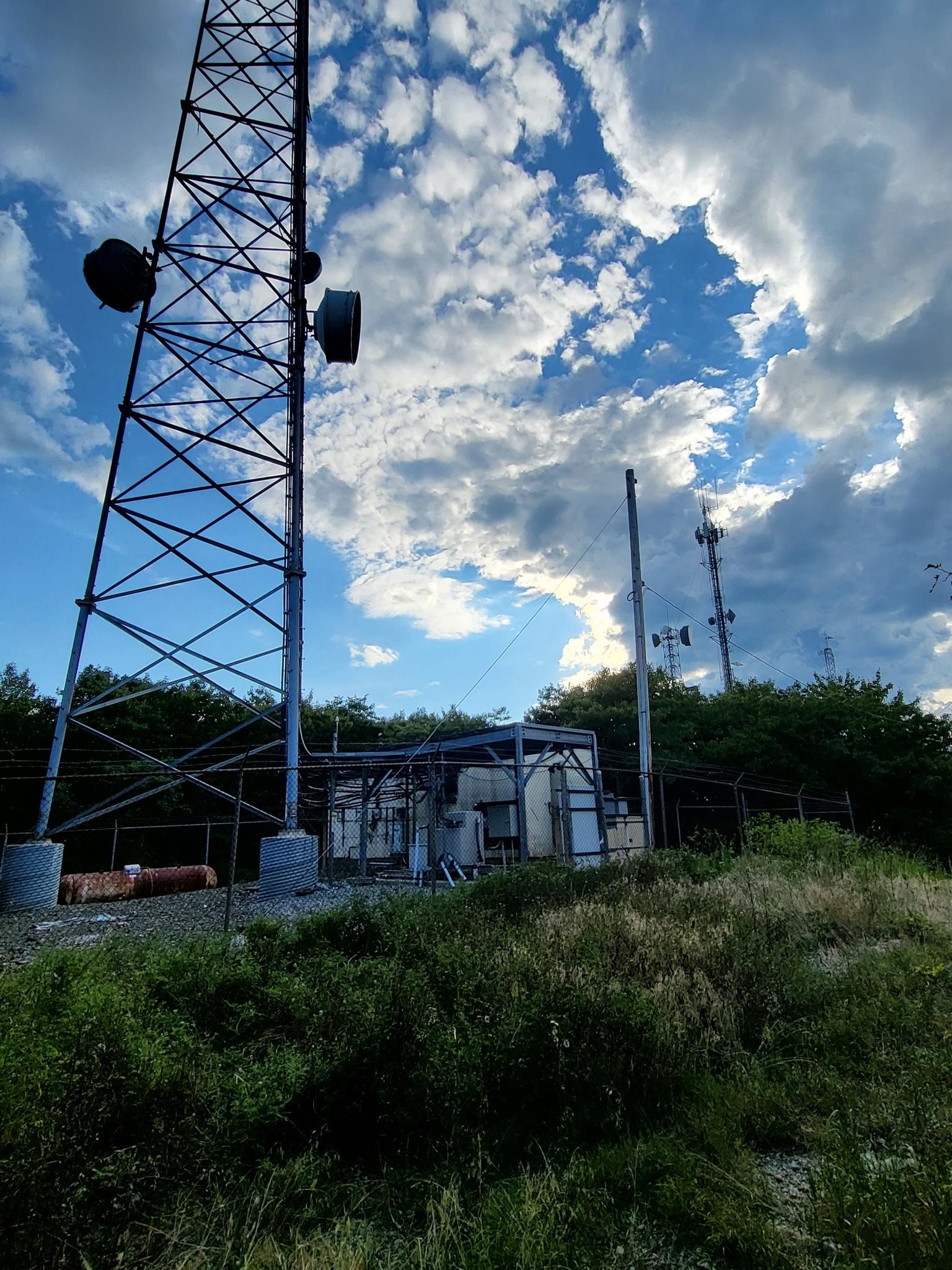 Tall communication towers stand against a dramatic cloudy sky on a grassy hill.