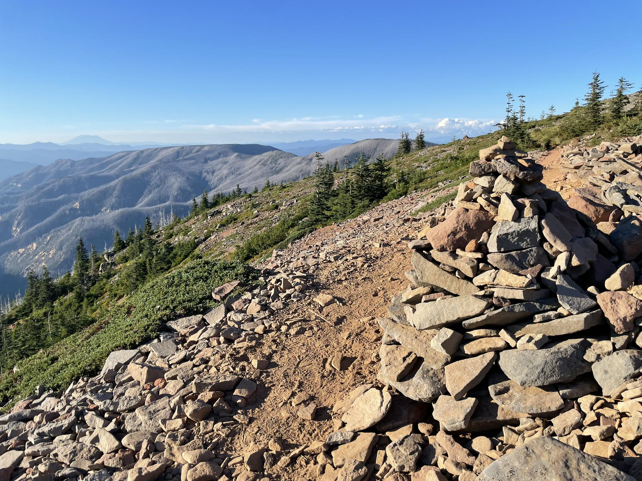A rock cairn marks a rocky mountain ridge with distant peaks under a clear sky.
