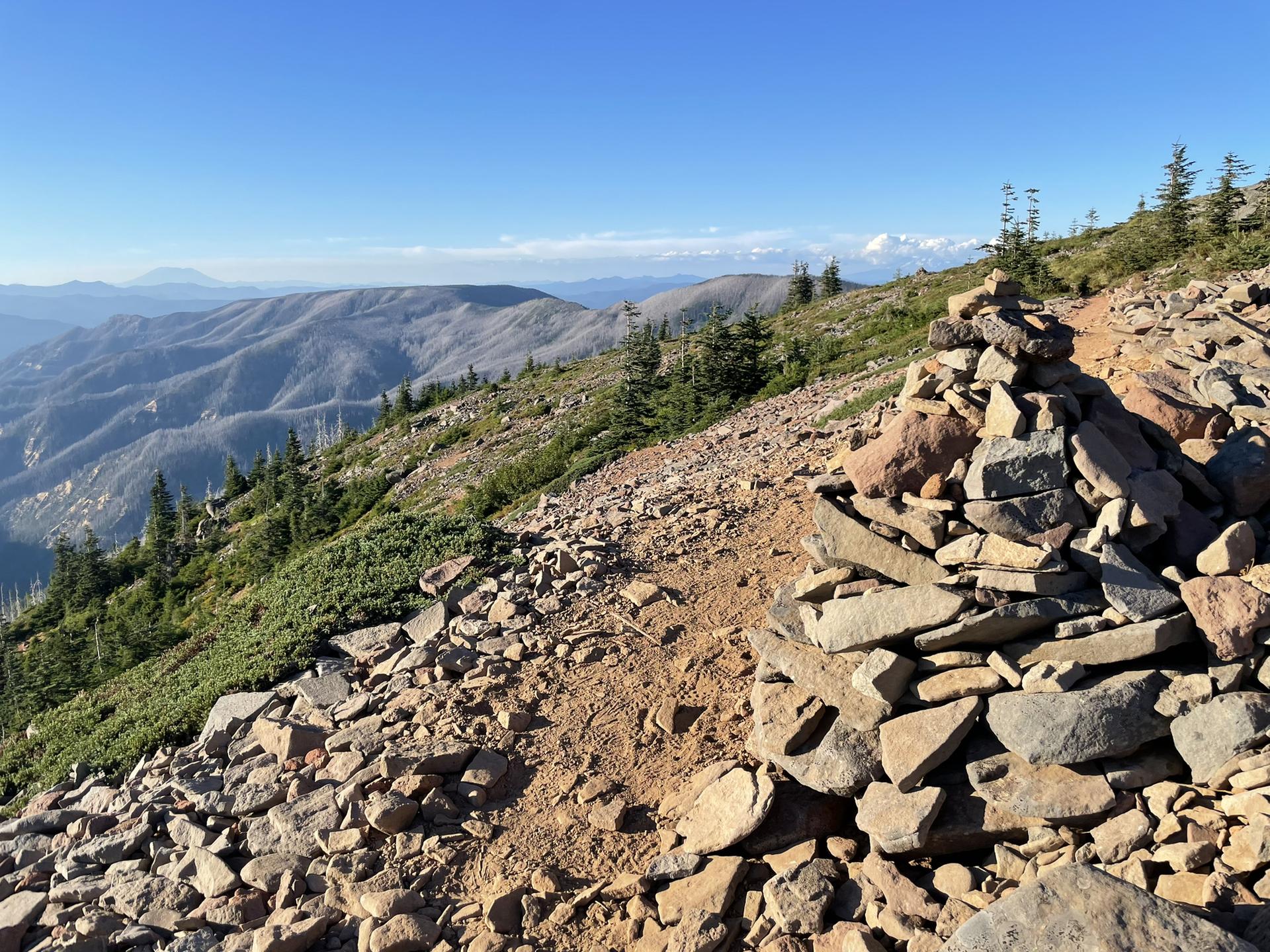 A rock cairn marks a rocky mountain ridge with distant peaks under a clear sky.