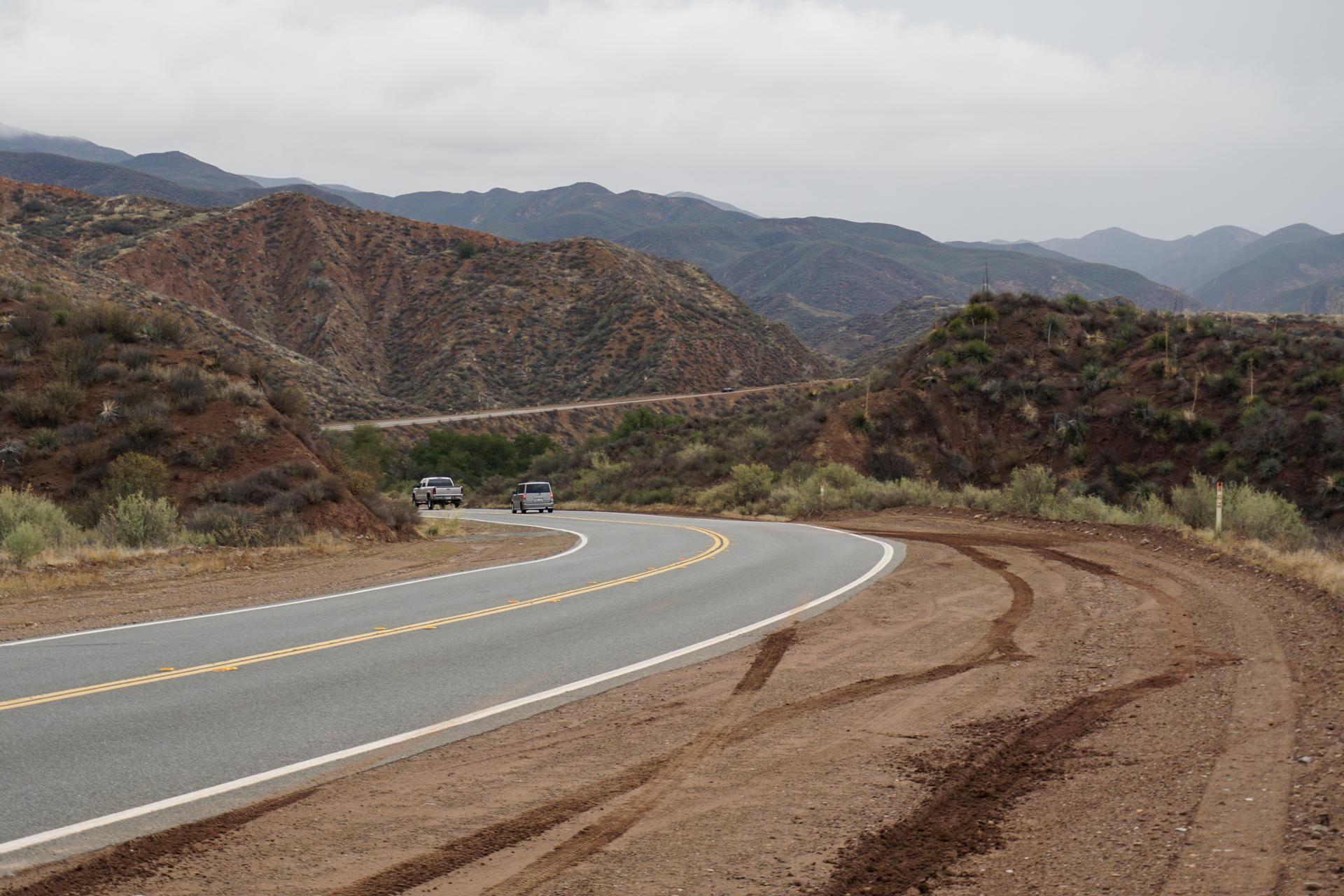 Winding road with two cars, surrounded by arid mountains under an overcast sky.