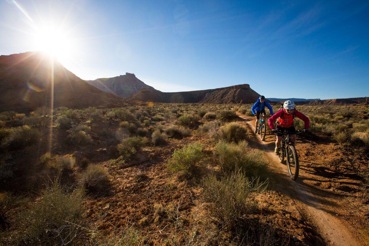 Two mountain bikers on a sunny desert trail with mountains.