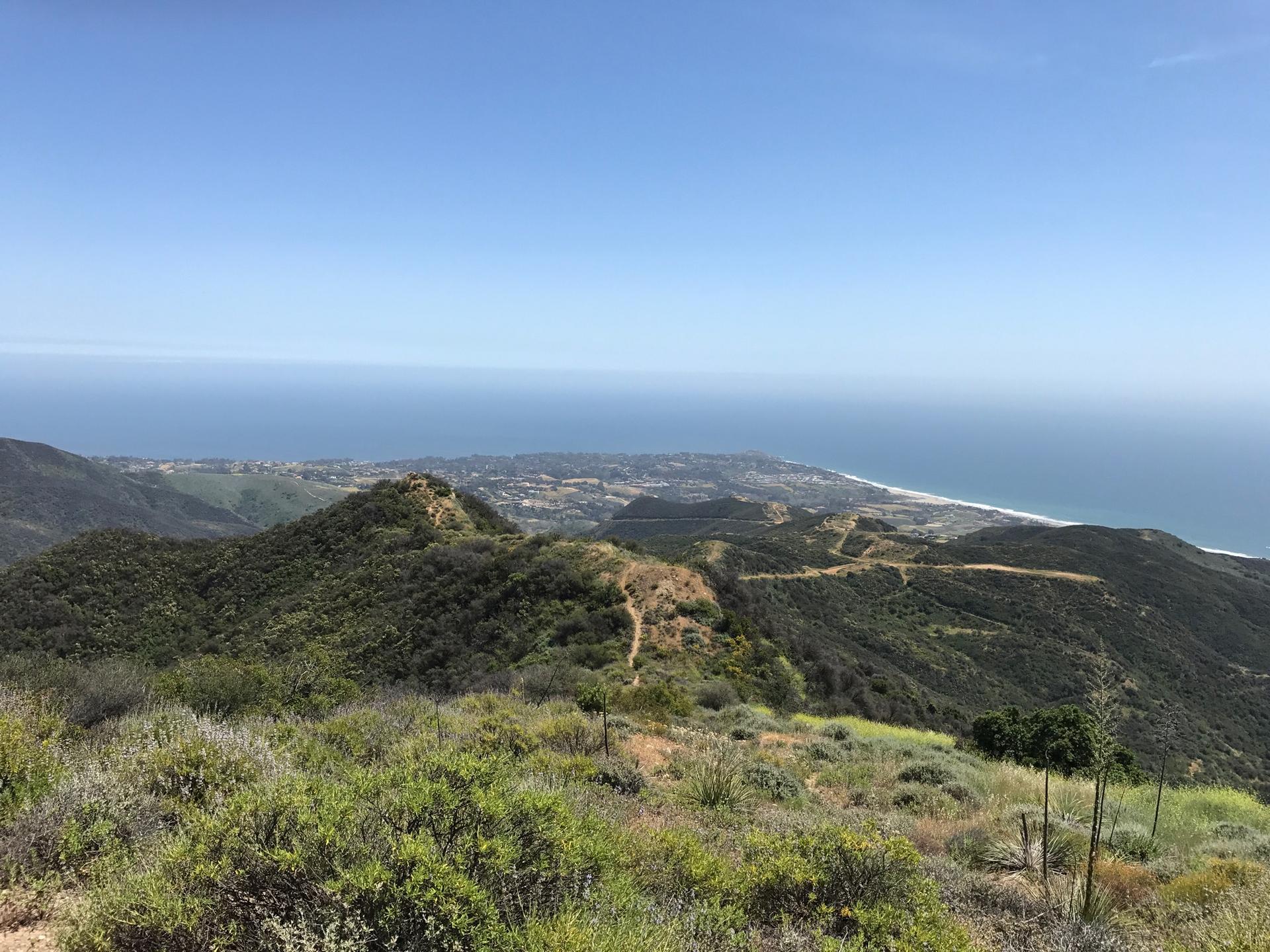 Panoramic view of green hillsides, distant coastline, and ocean under a clear sky.