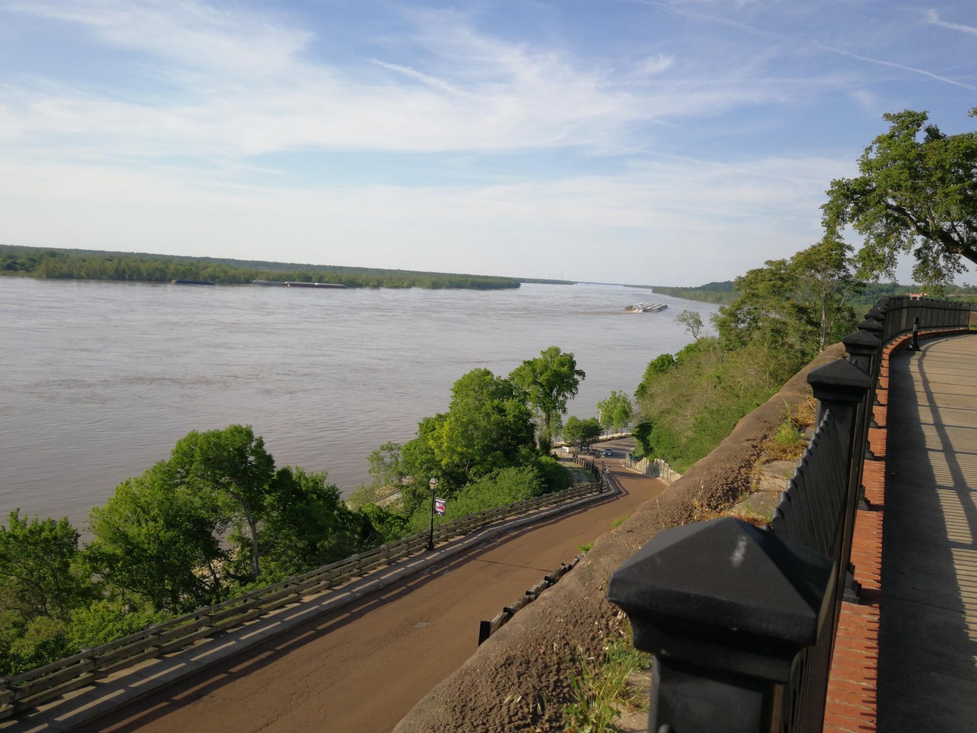 Wide river, curved path, and green trees under a bright, cloudy sky.