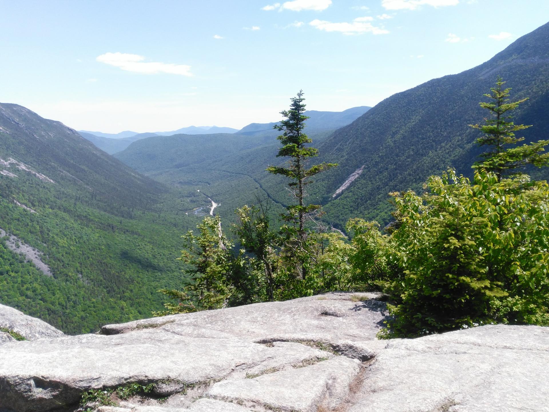 Rocky outcrop overlooking a green mountain valley under a clear sky.