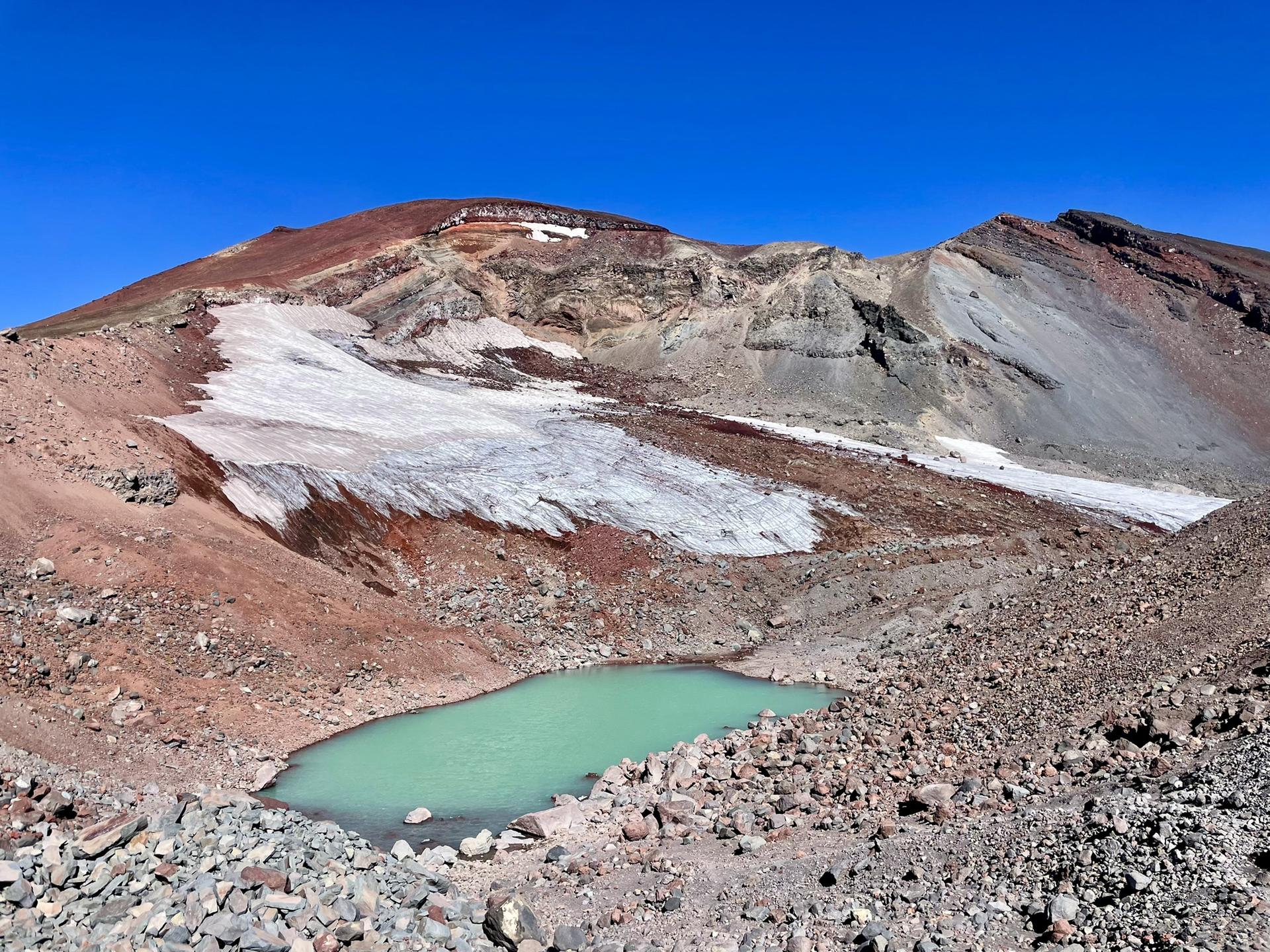 A small turquoise glacial lake at the base of a rocky, snow-covered mountain.