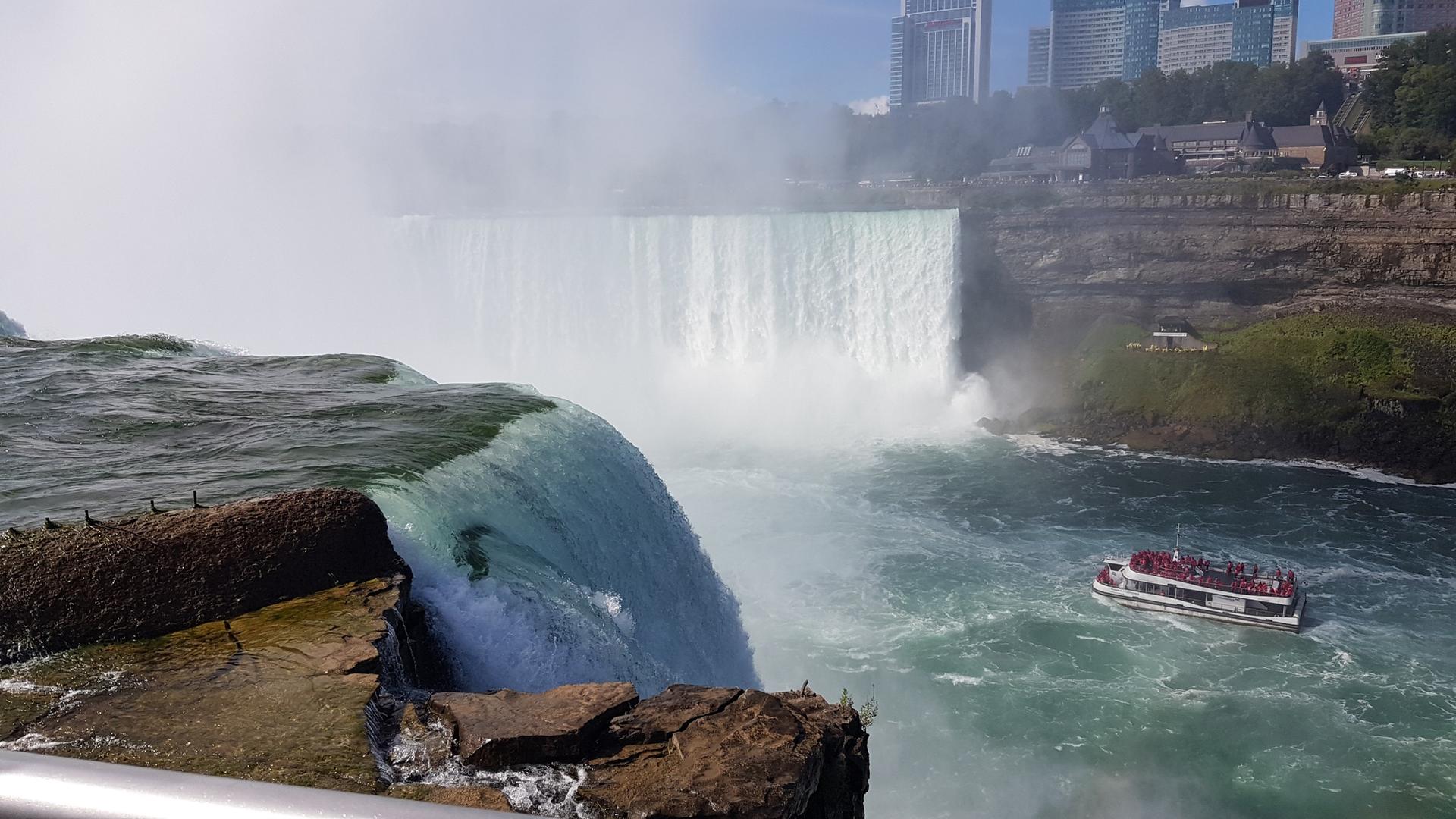 Powerful waterfall with mist, a boat, and city buildings on a bright day.