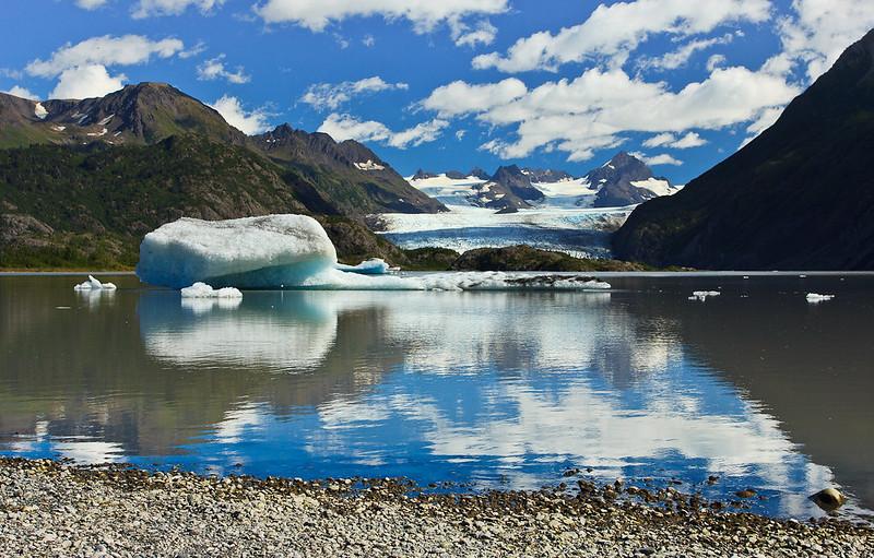 Bright sunny day with a large iceberg in a lake, mountains, and glacier.