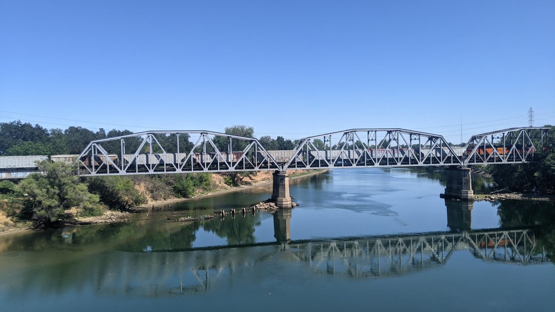 A long metal truss bridge crosses a calm river under a bright blue sky.